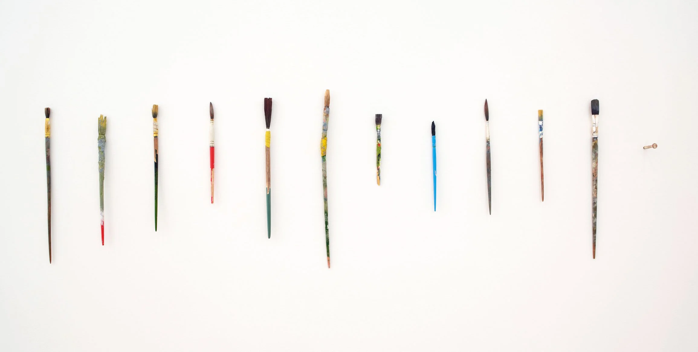 A row of small artist paintbrushes with colorful paint residue on the bristles and handles, arranged in a line on a white background.