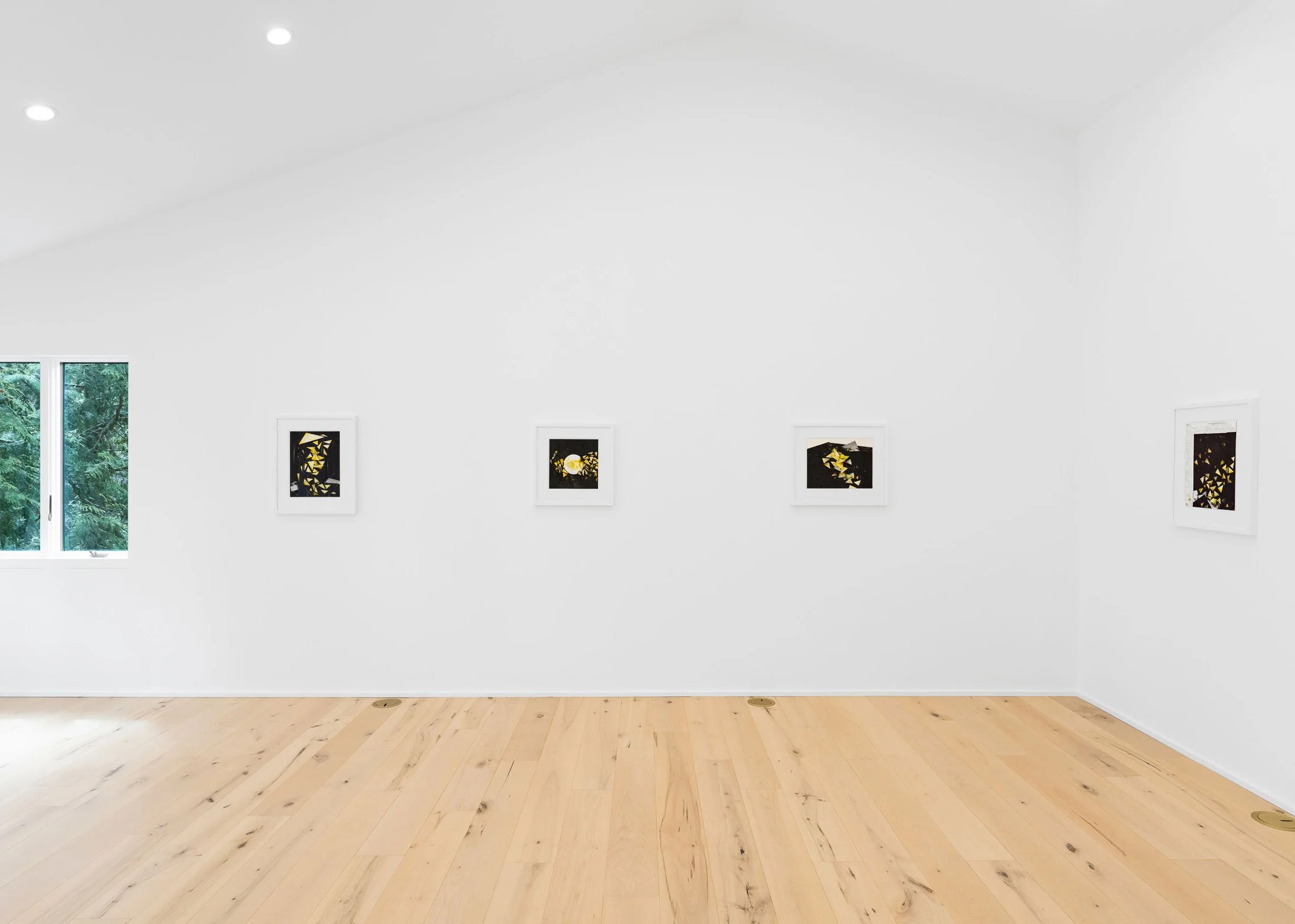 An art gallery with three abstract framed artworks on a white wall, a window showing lush green trees outside, and a light hardwood floor.