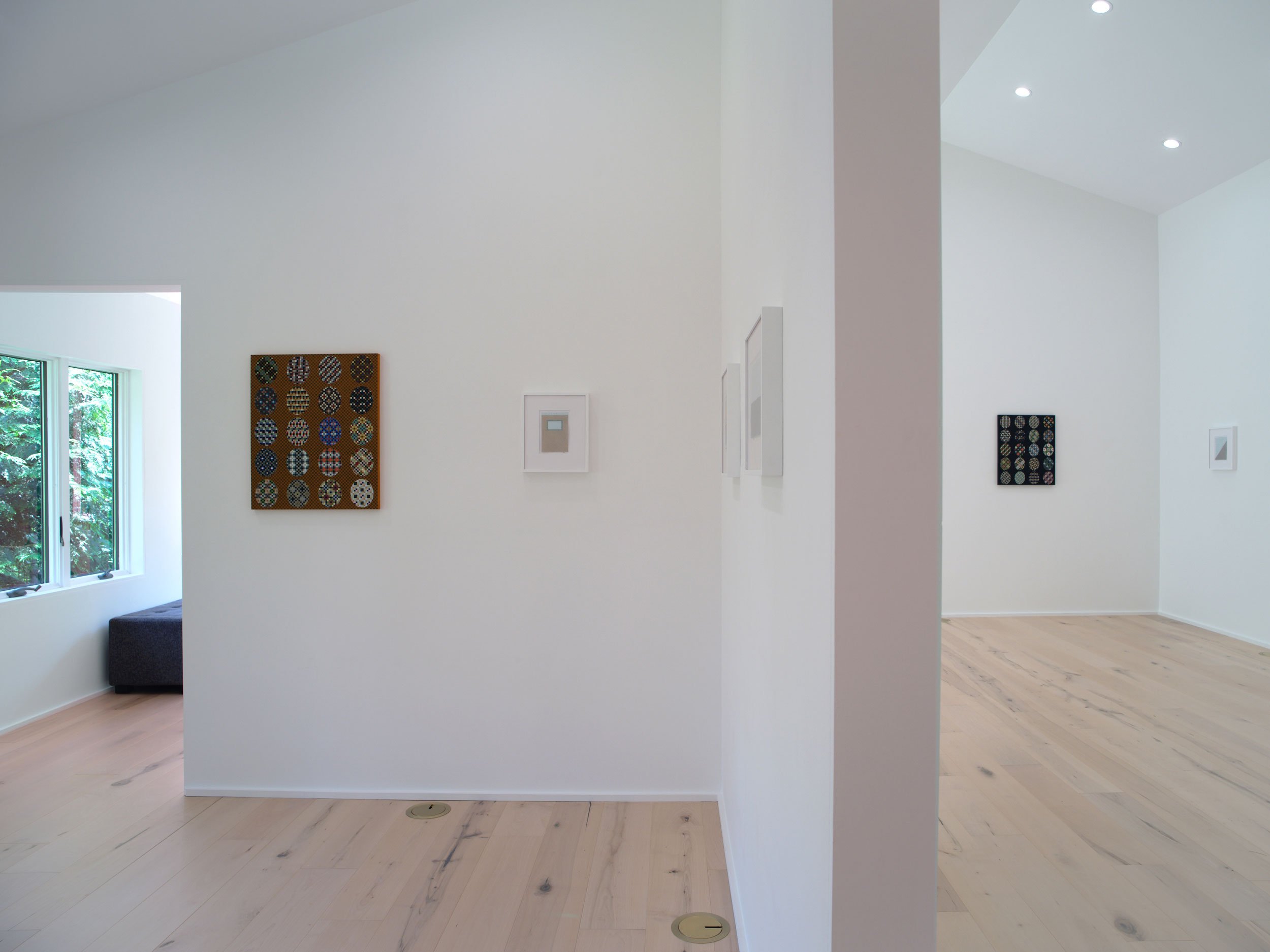 A minimalist interior of a bright room with white walls and wooden flooring. Artworks hang on the walls, and a window shows greenery outside. There are recessed ceiling lights.