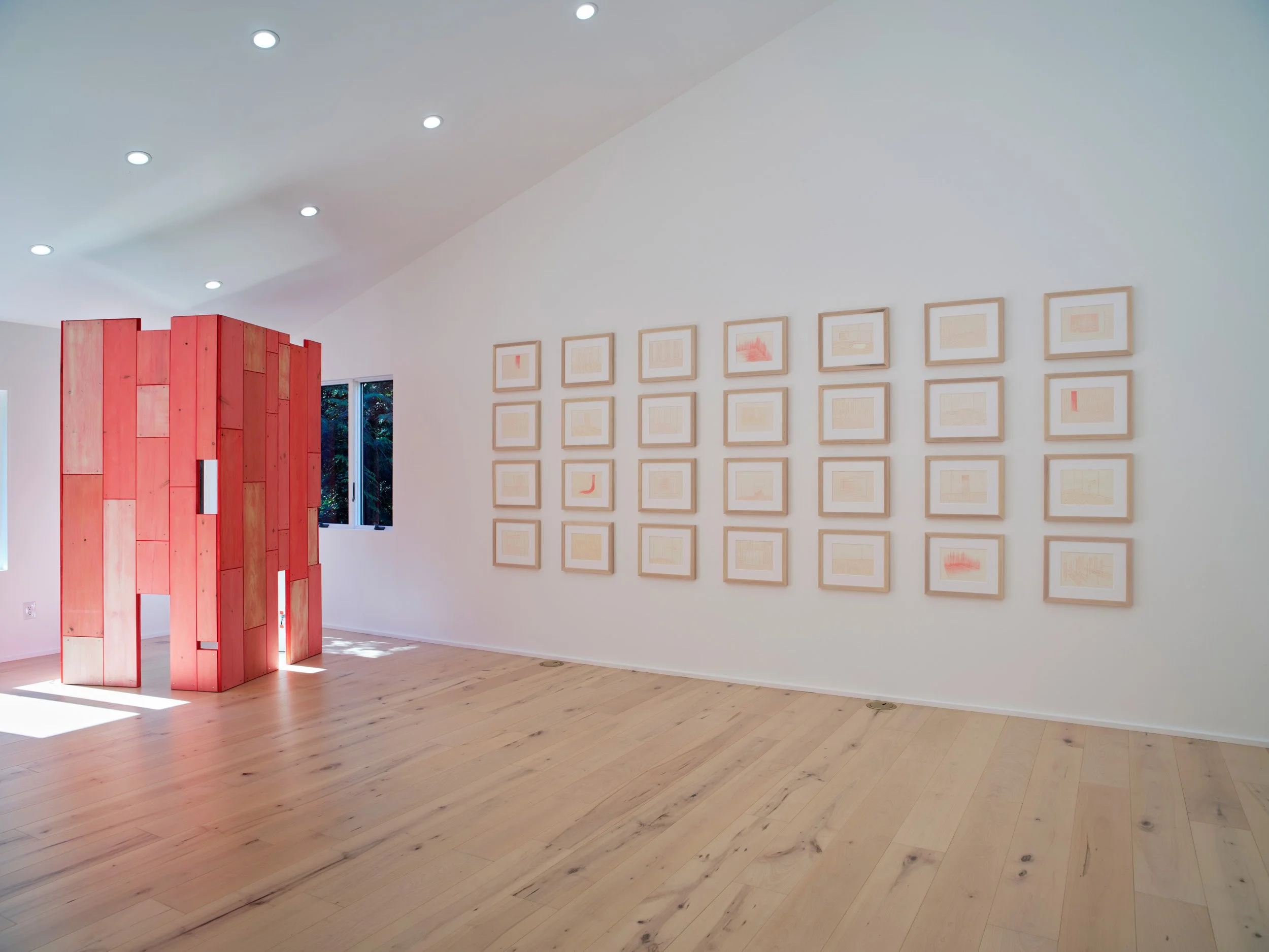 Interior view of an art gallery with a white wall featuring a grid of framed abstract artwork and a curved red wooden room divider, with hardwood flooring and ceiling lights.