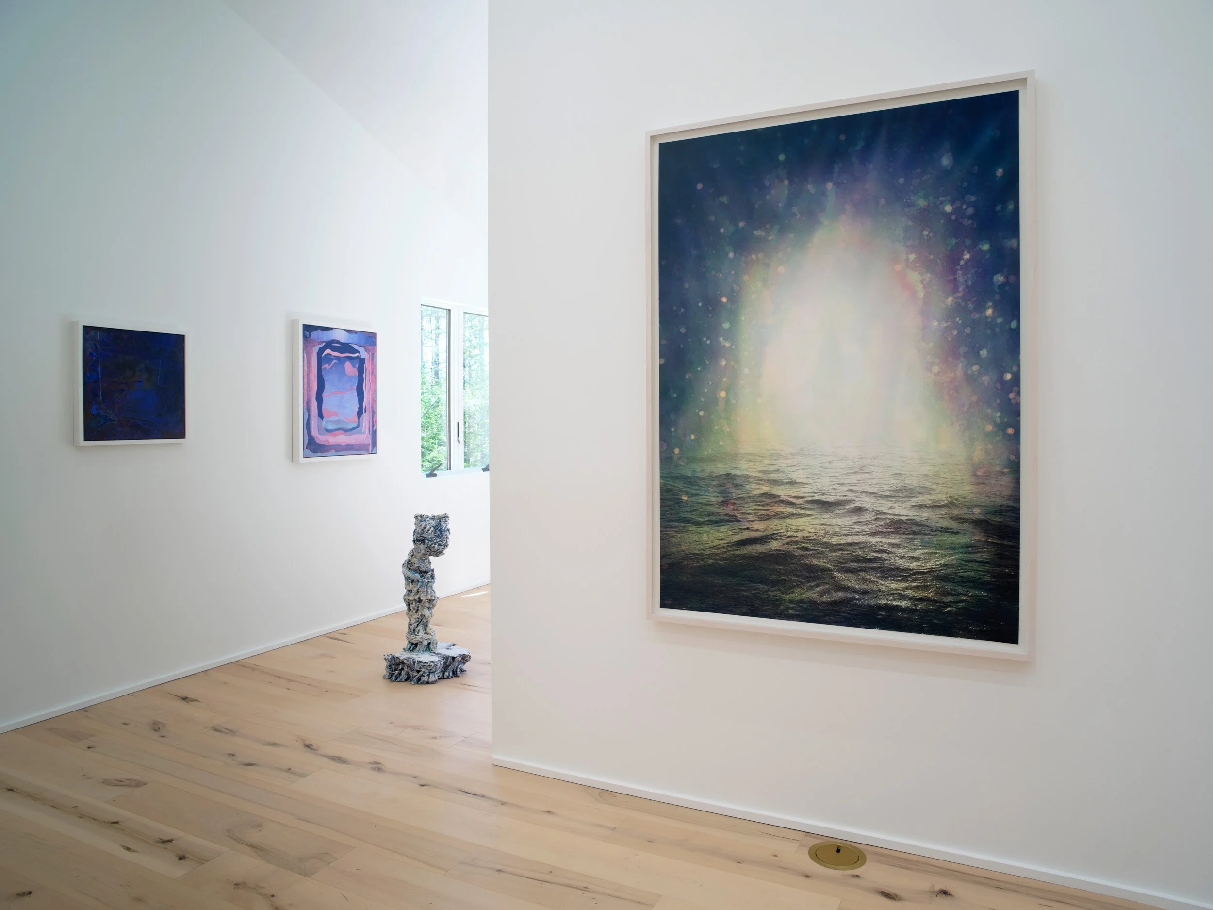 An art gallery with three framed paintings on white walls and a sculpture in front. The largest painting depicts an abstract scene of water with an iridescent light, and the other two are smaller with abstract designs. There is a window in the background letting in natural light, and the gallery has light wood floors.