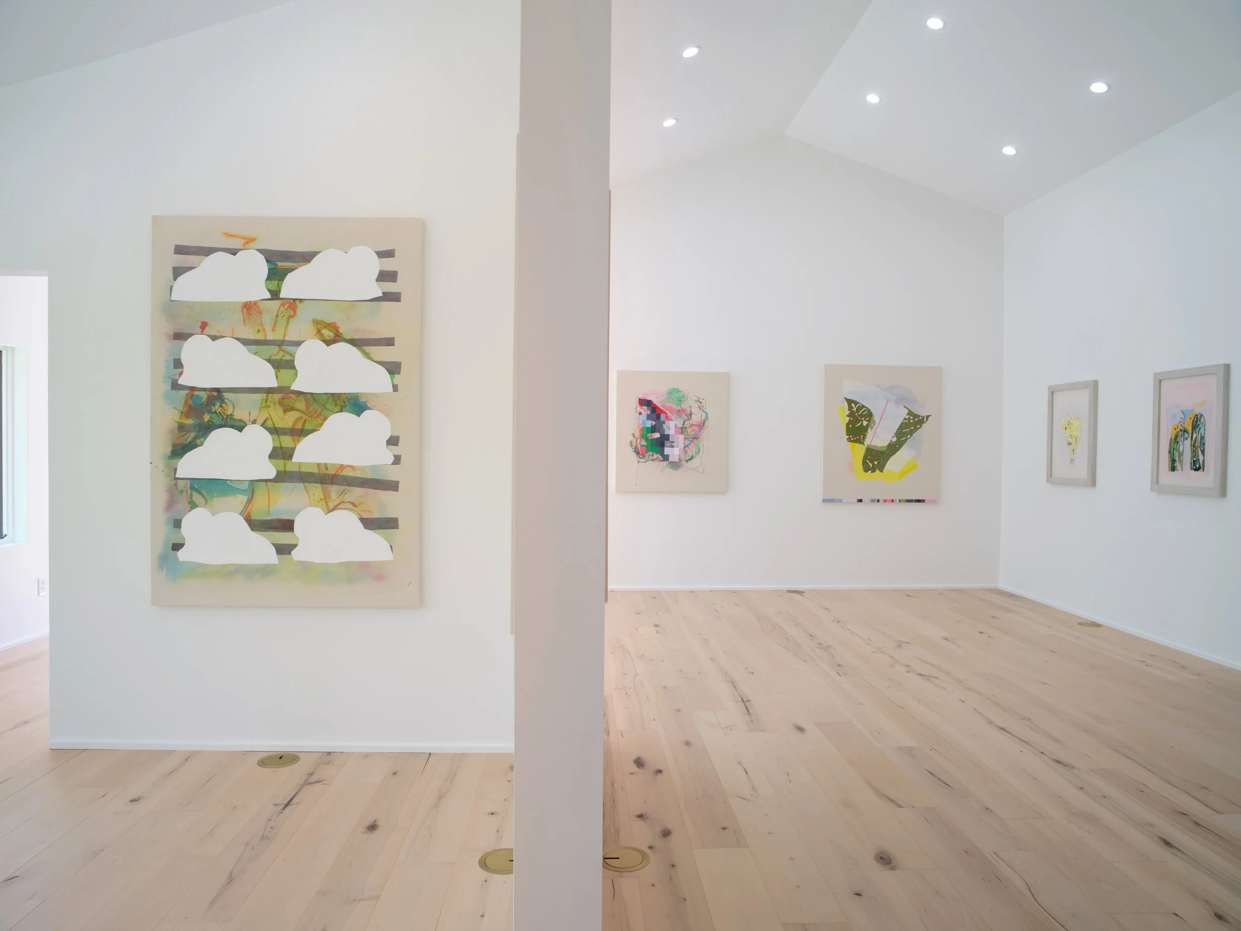 An art gallery with white walls displaying colorful modern art. The image is split by a white column in the foreground, with ceiling lights illuminating the artwork.