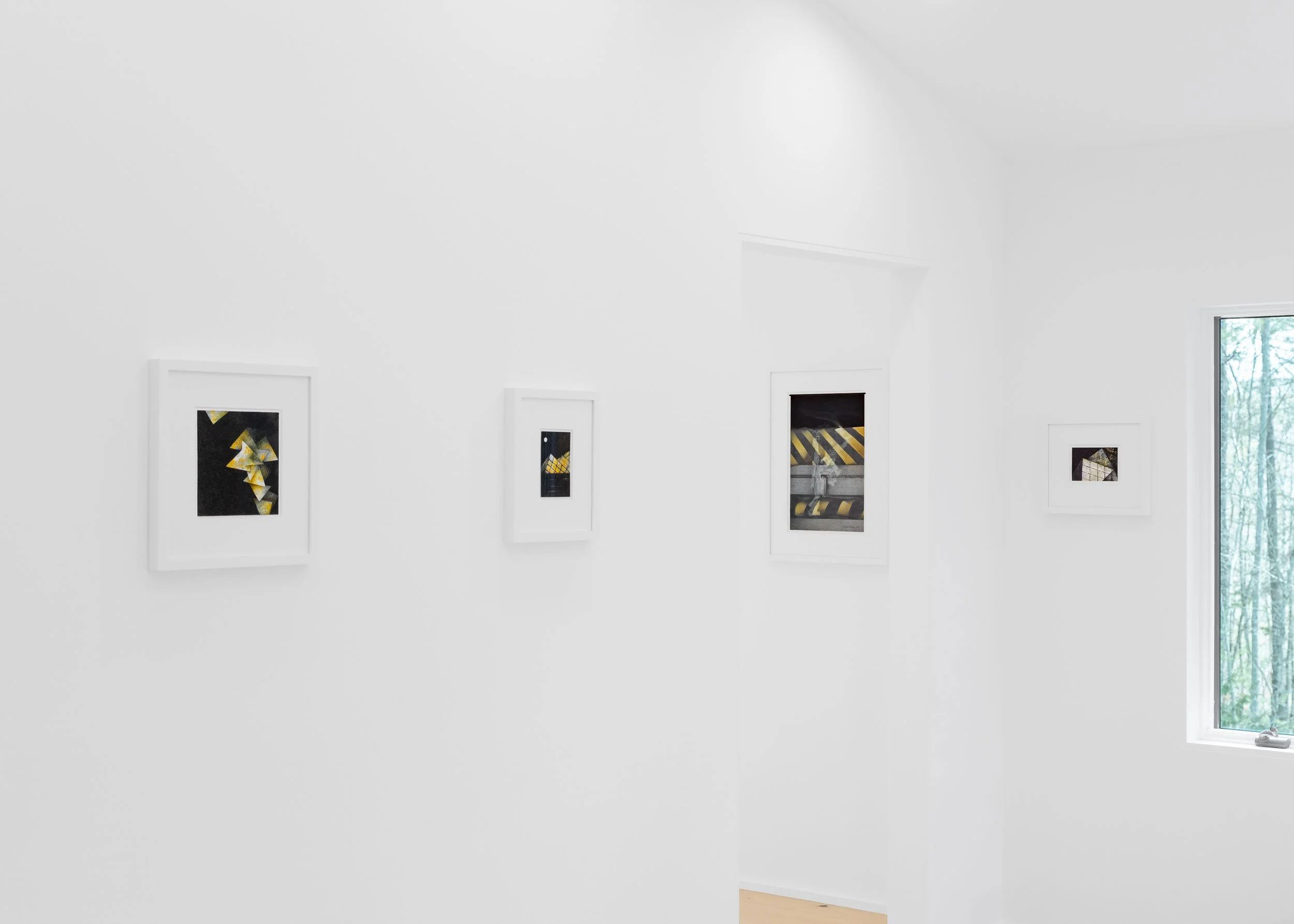 Interior of a white art gallery with five framed artworks on the wall and a window showing trees outside.