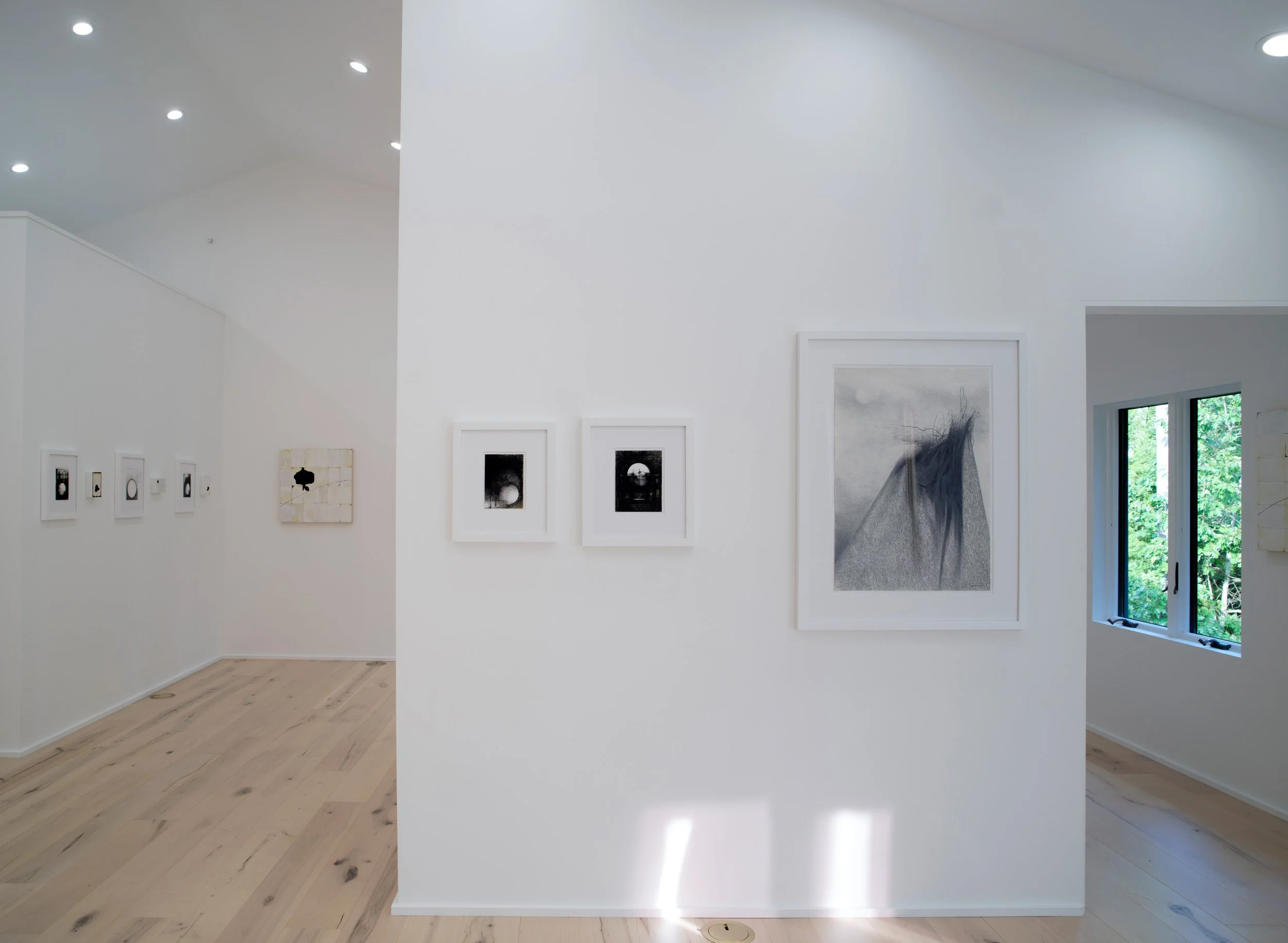 Interior of an art gallery with white walls and framed artwork, including black and white photographs and abstract pieces, natural light from a window, and wooden flooring.