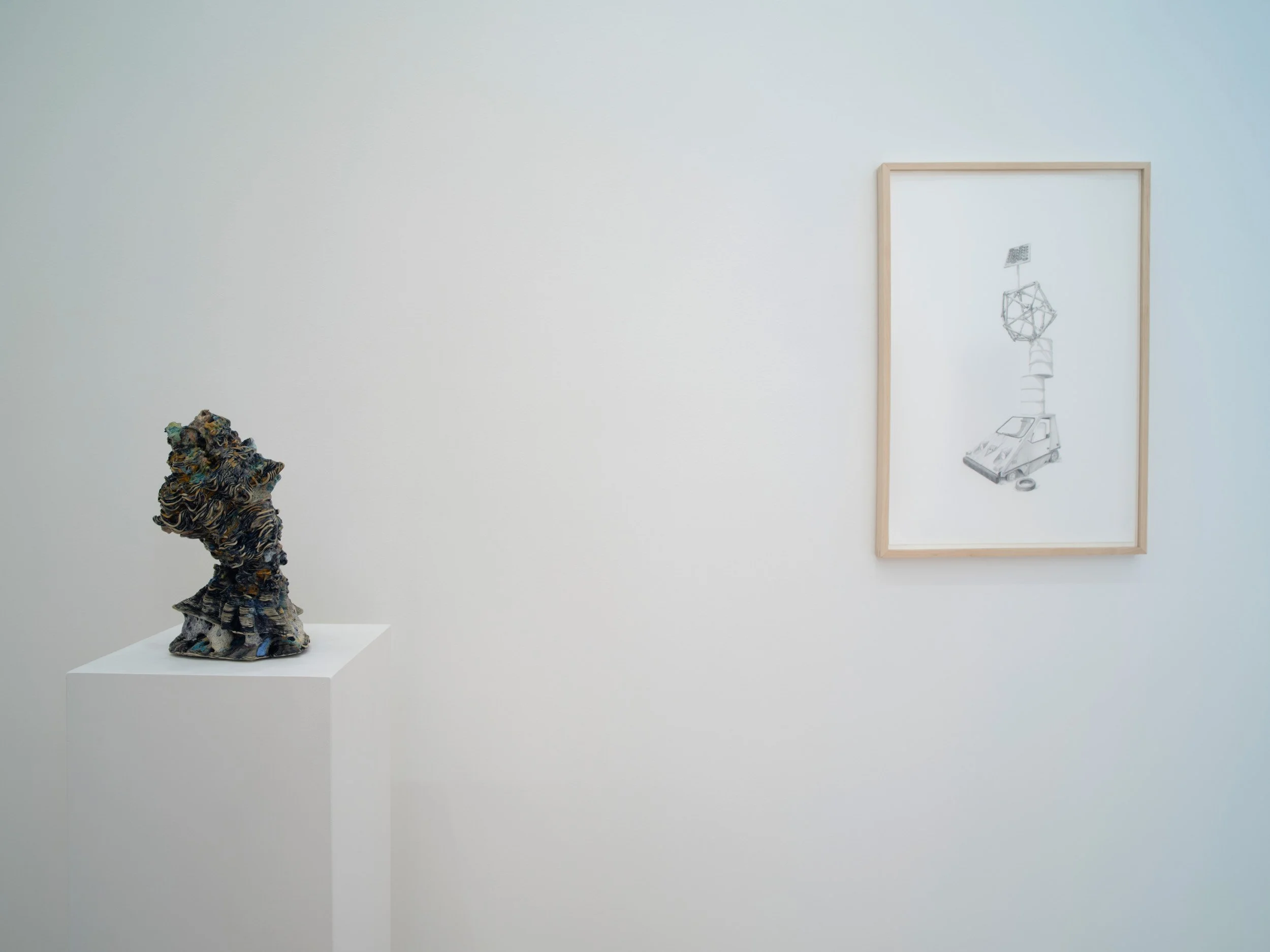 An art gallery with a abstract sculpture on a pedestal to the left and a framed sketch of a vehicle with a geometric object on top hanging on the wall to the right.