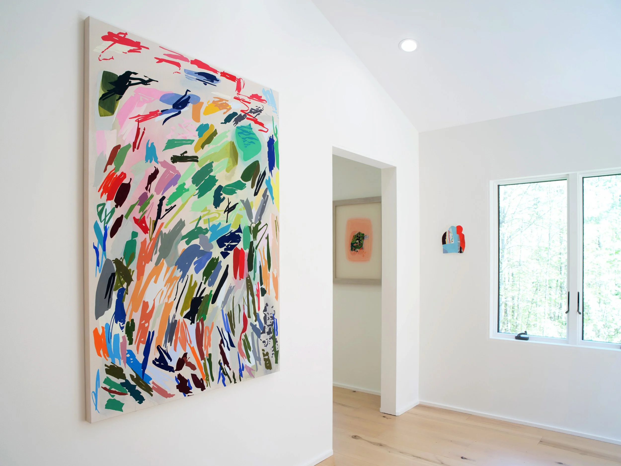 An art gallery with abstract paintings on white walls, natural light coming through windows, and light-colored wooden flooring.