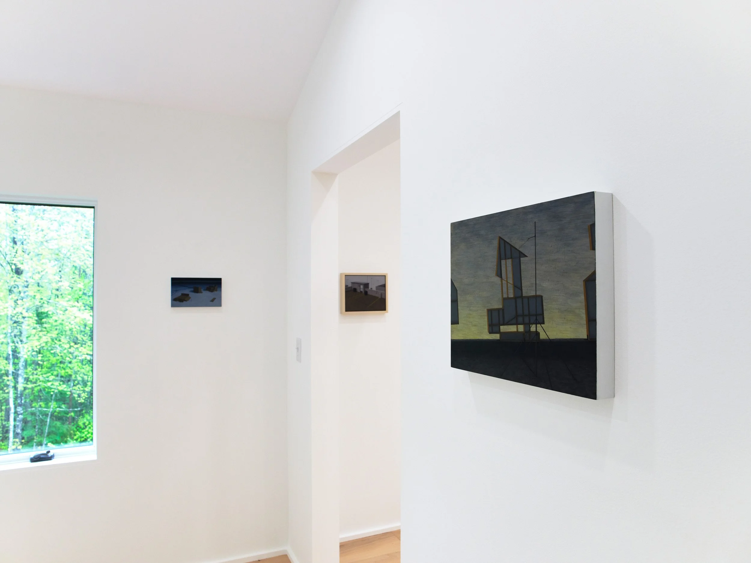 Interior view of a modern art gallery with white walls, featuring several framed artworks. There is a large window on the left side showing green trees outside.