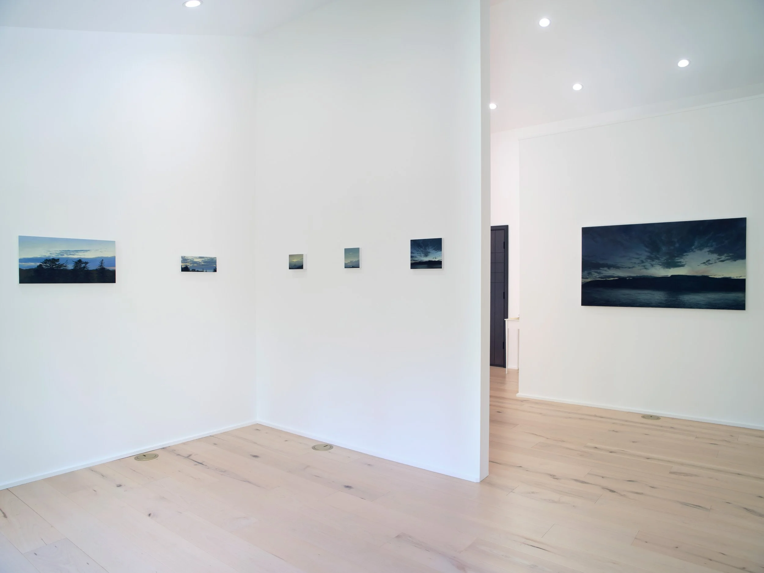 A white-walled art gallery with landscape paintings hung on the walls, featuring views of the sky and nature. The gallery has light wooden flooring and ceiling spotlights.