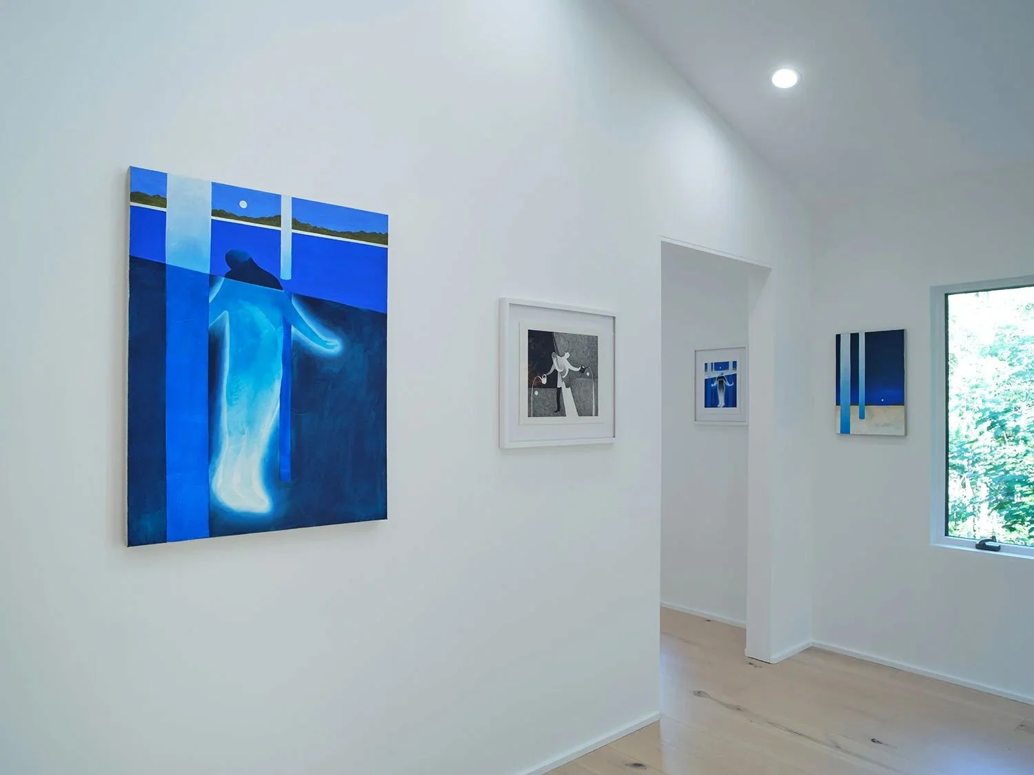 Art gallery with multiple paintings on white walls, featuring various abstract and figurative artworks, some in blue and monochrome tones, near a window with greenery outside.
