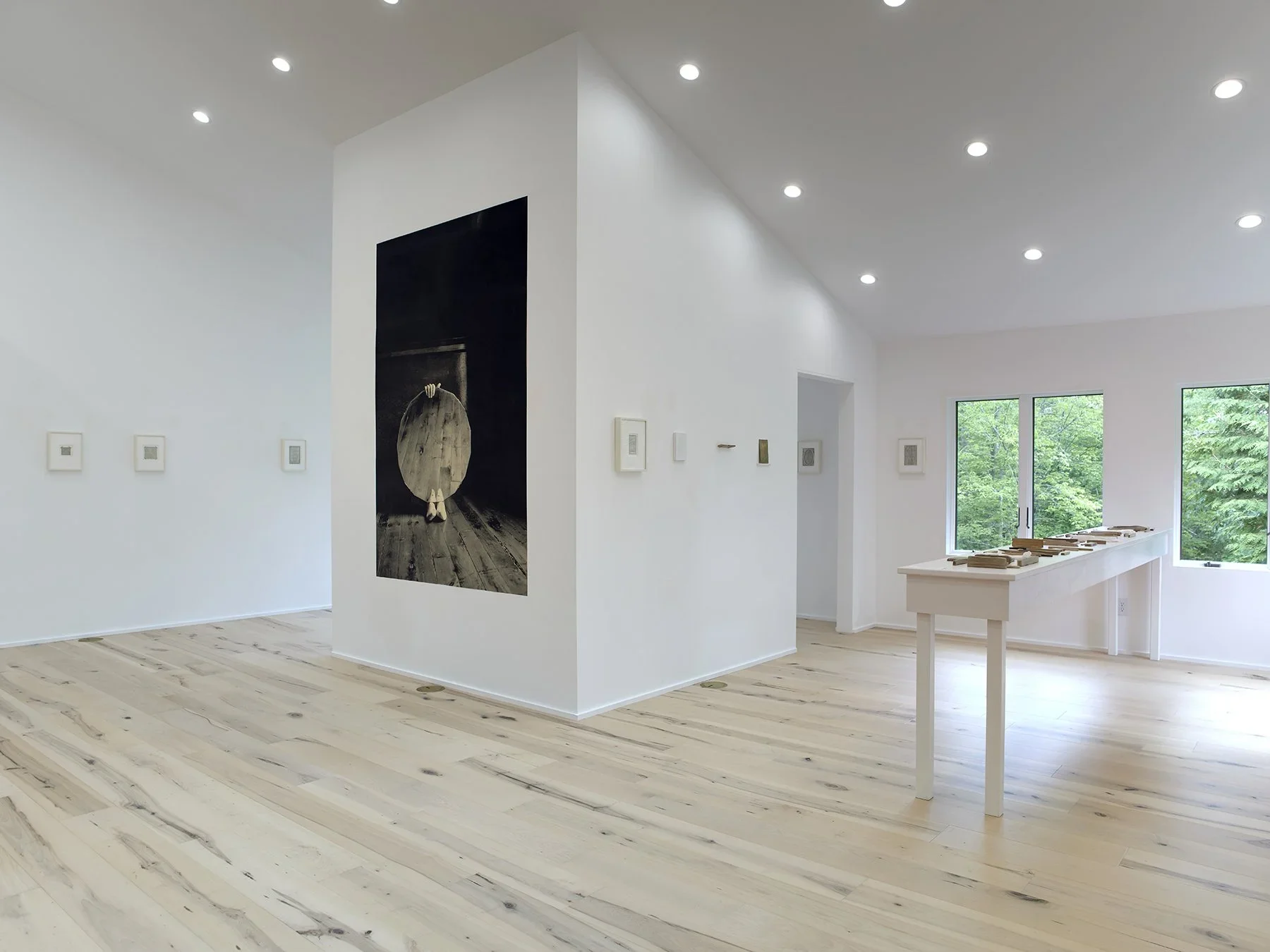 An art gallery with white walls and light wooden floors, featuring several framed artworks and a large black-and-white photograph of a girl with a large barrel.