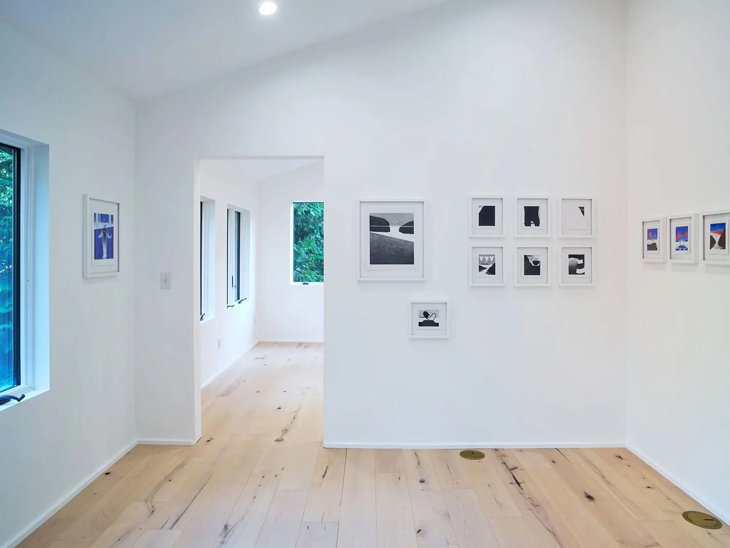 Art gallery with white walls and multiple framed photographs or artworks arranged on the walls, natural light coming through windows, light wood flooring.