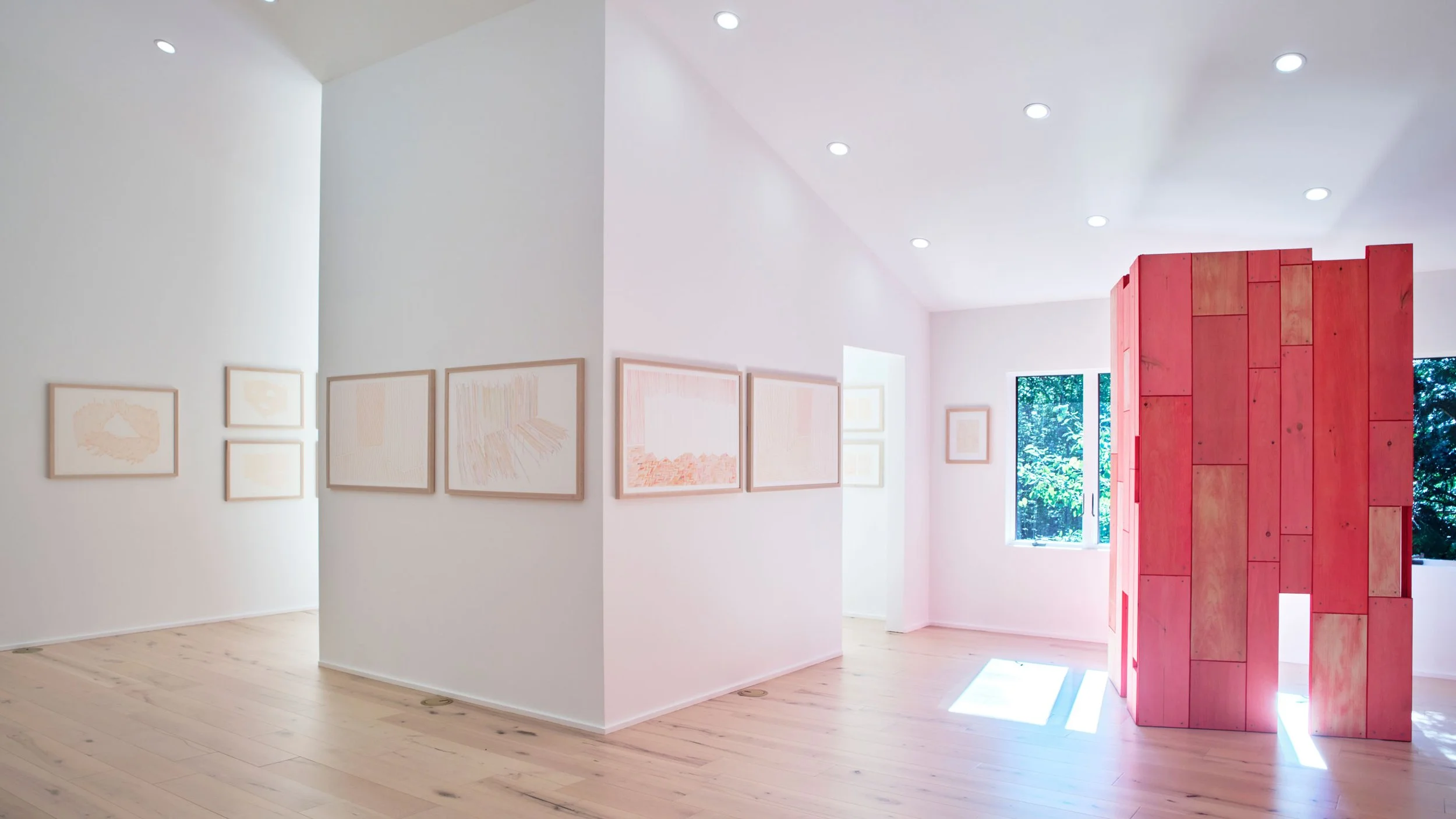 Art gallery with white walls, framed artworks on the walls, a large pink wooden structure, wooden floors, and windows allowing natural light