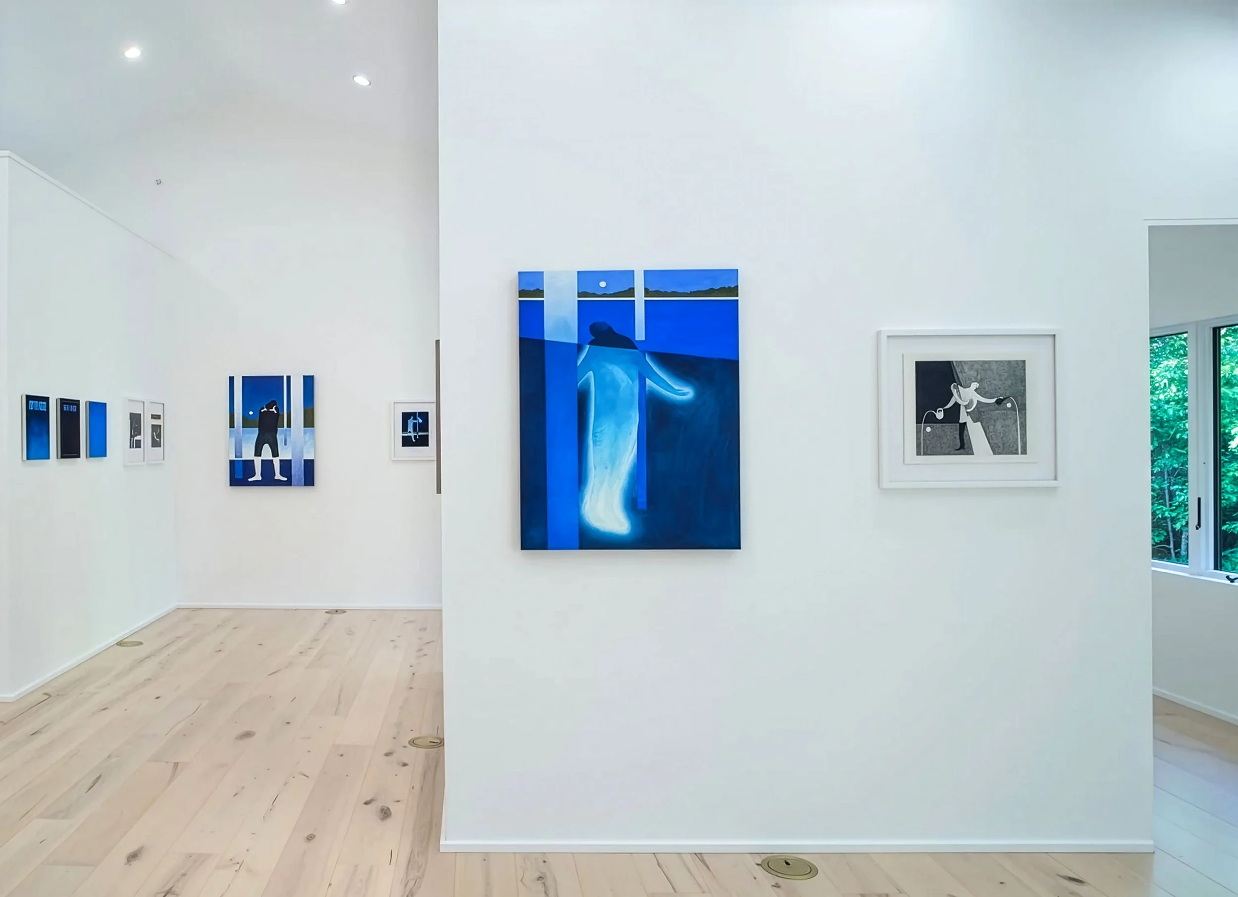 White-walled art gallery with several paintings, mostly blue and black, hung on the walls. Large windows on the right side showing greenery outside.