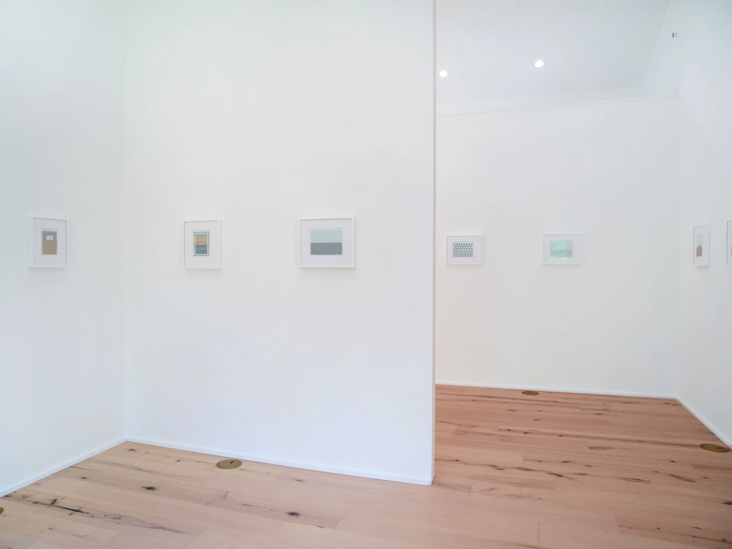 Gallery view of framed artwork on white walls, contemporary art pieces, wooden flooring, and minimalistic display in an art gallery.