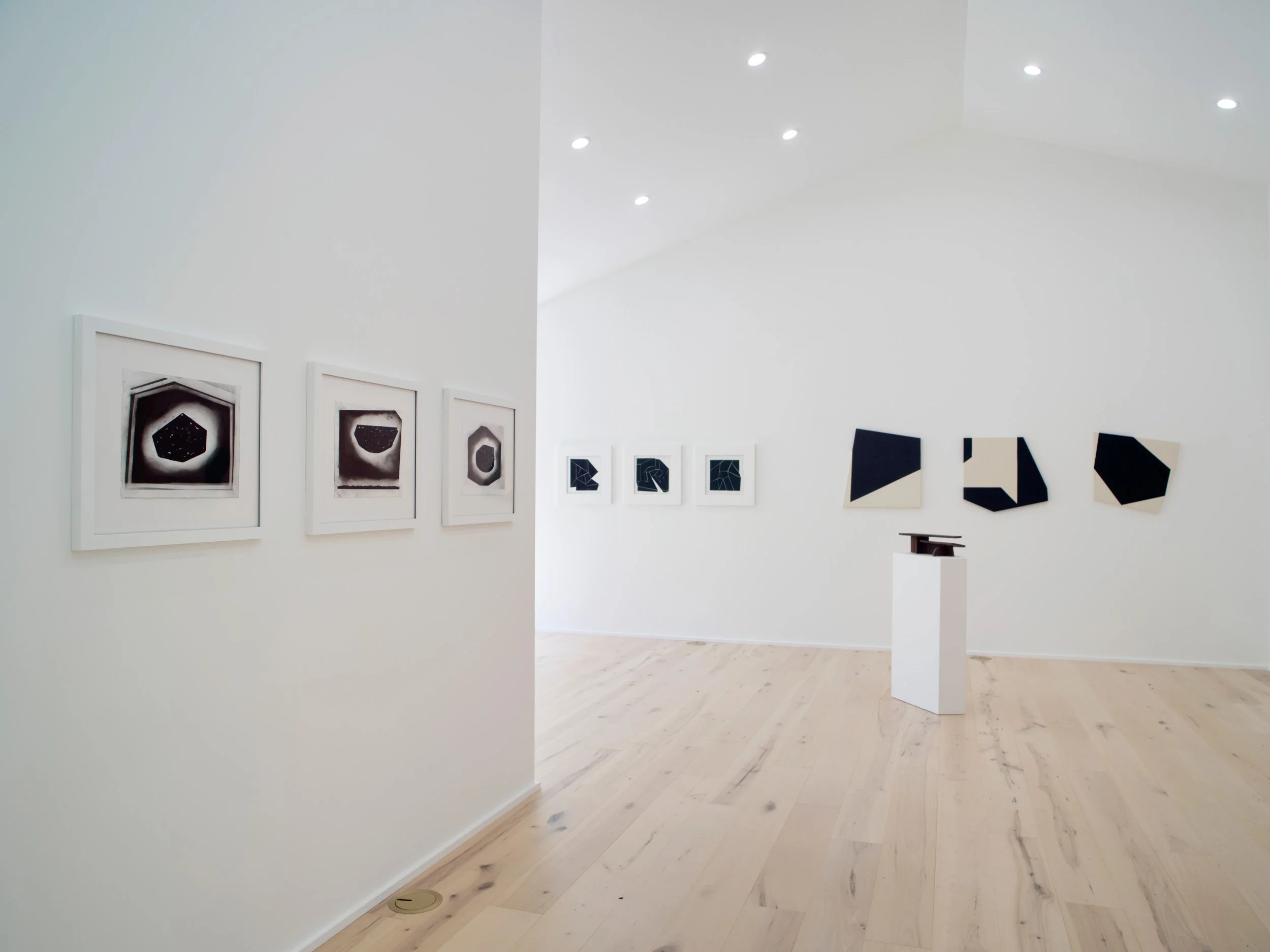An art gallery with white walls displaying framed black and white abstract artwork and geometric paintings, wooden flooring, and bright ceiling lights.