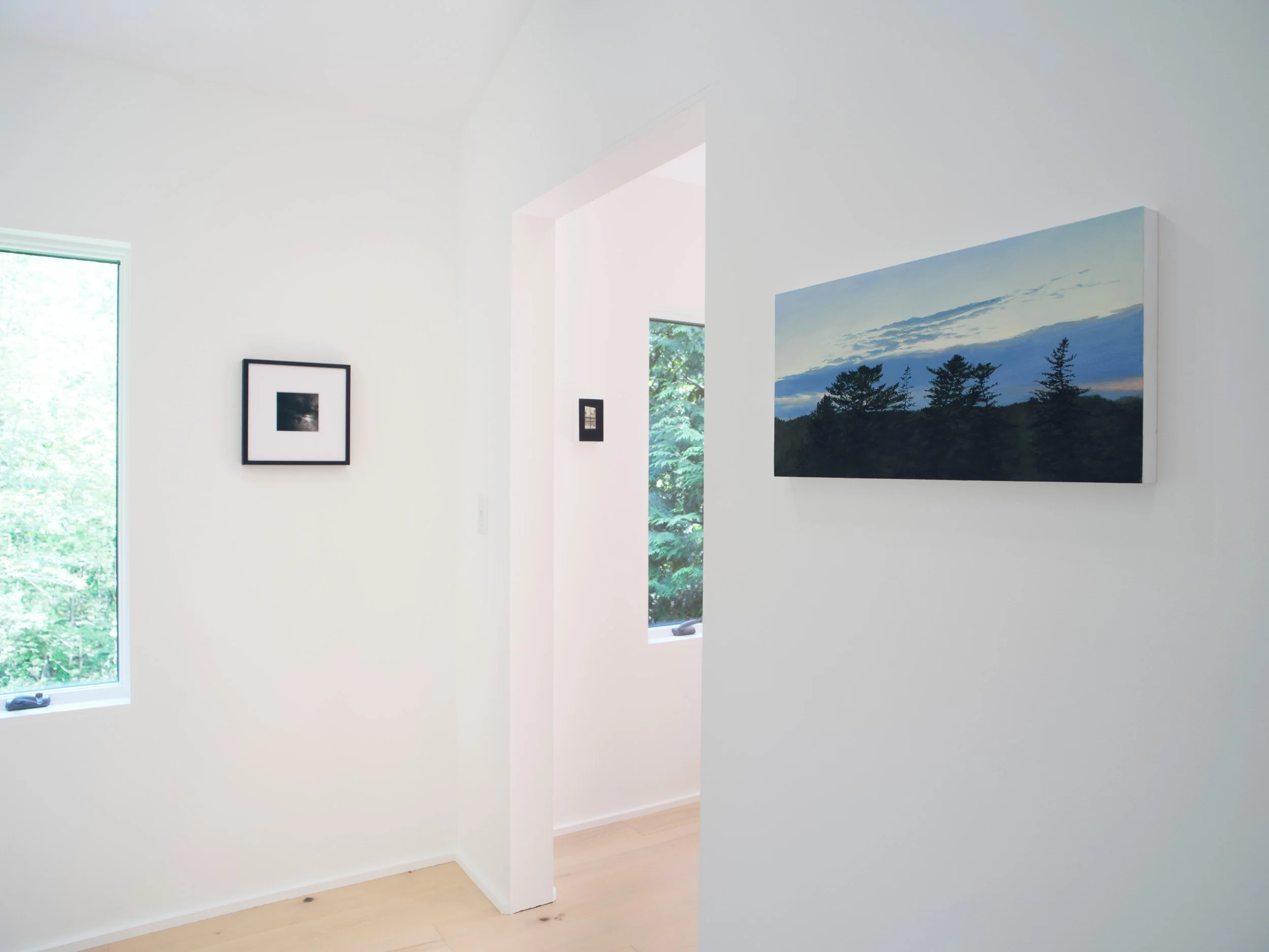 Interior view of a room with white walls, two windows letting in natural light, and framed pictures on the wall, including a large landscape painting.