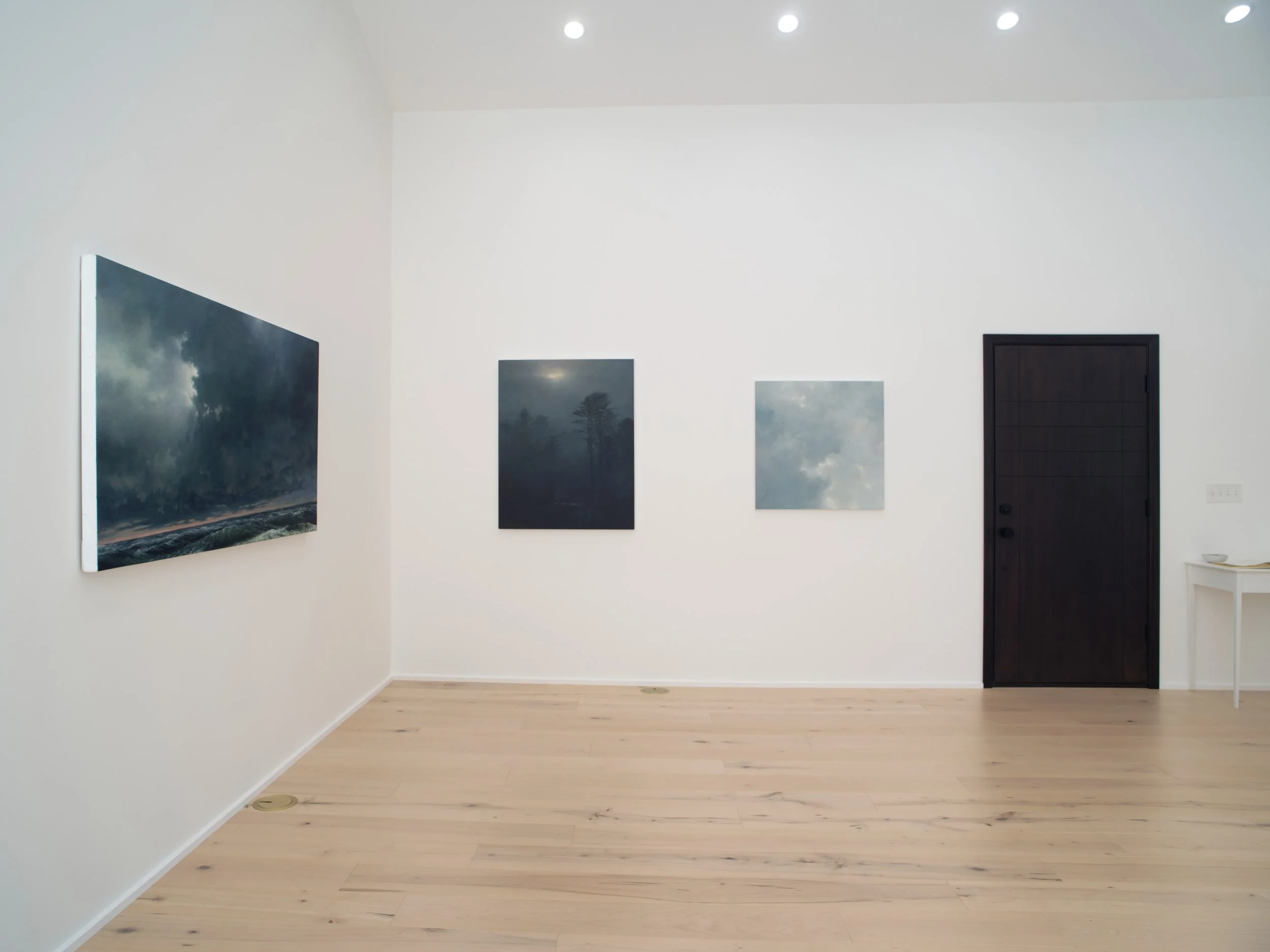 An art gallery with three dark cloudy landscape paintings on a white wall, a black door, and a small white table in the corner.