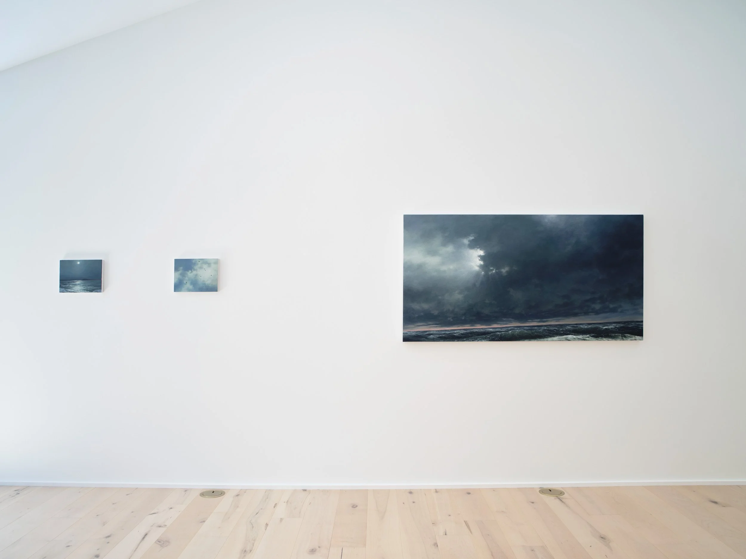 Gallery wall featuring four photographs of stormy skies with dark clouds in an art gallery with white walls and light wooden flooring.