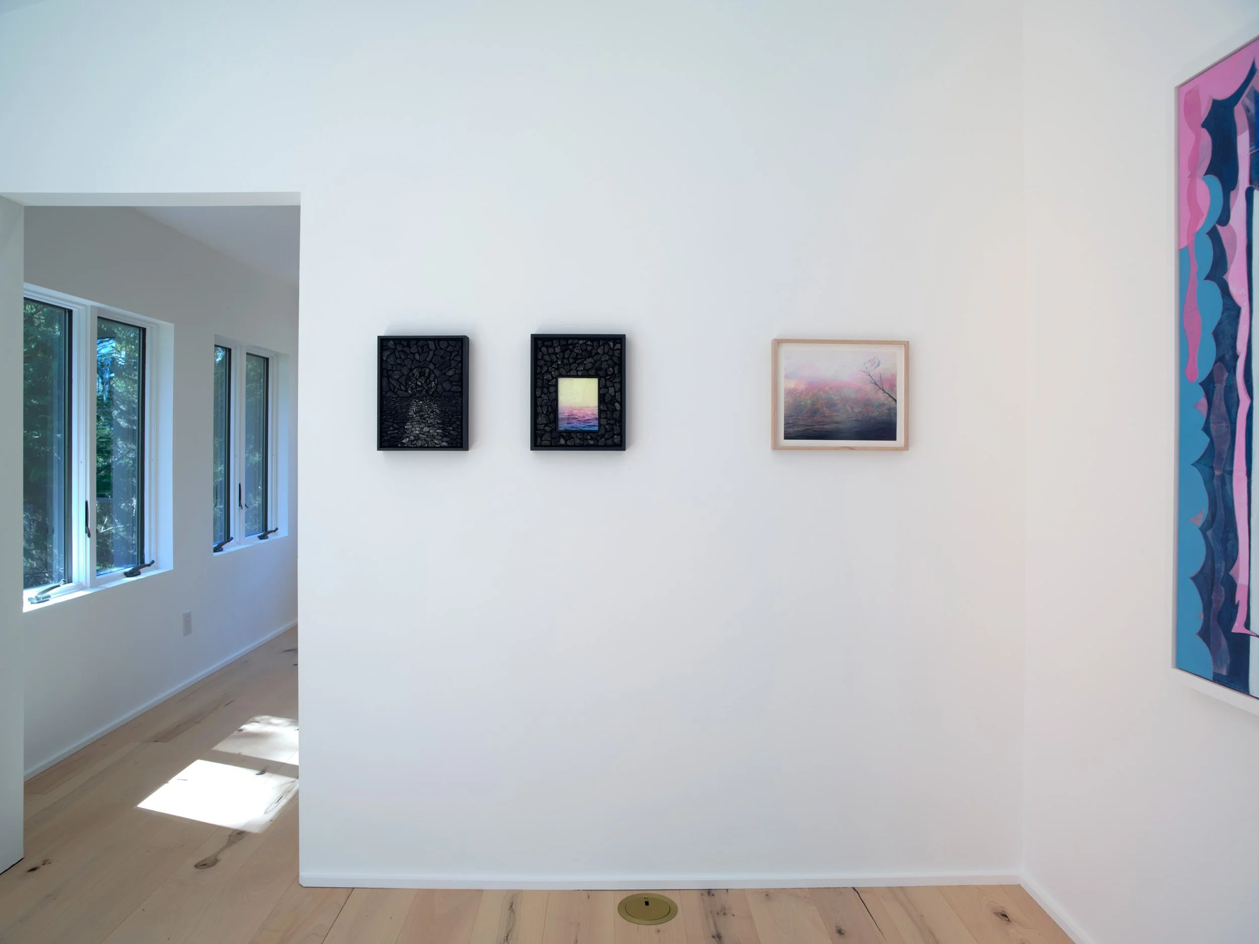 An art gallery with white walls displaying various paintings, including two framed pieces with black backgrounds and a pink forest scene, and a large, vertical, abstract painting with pink and blue shapes on the right.
