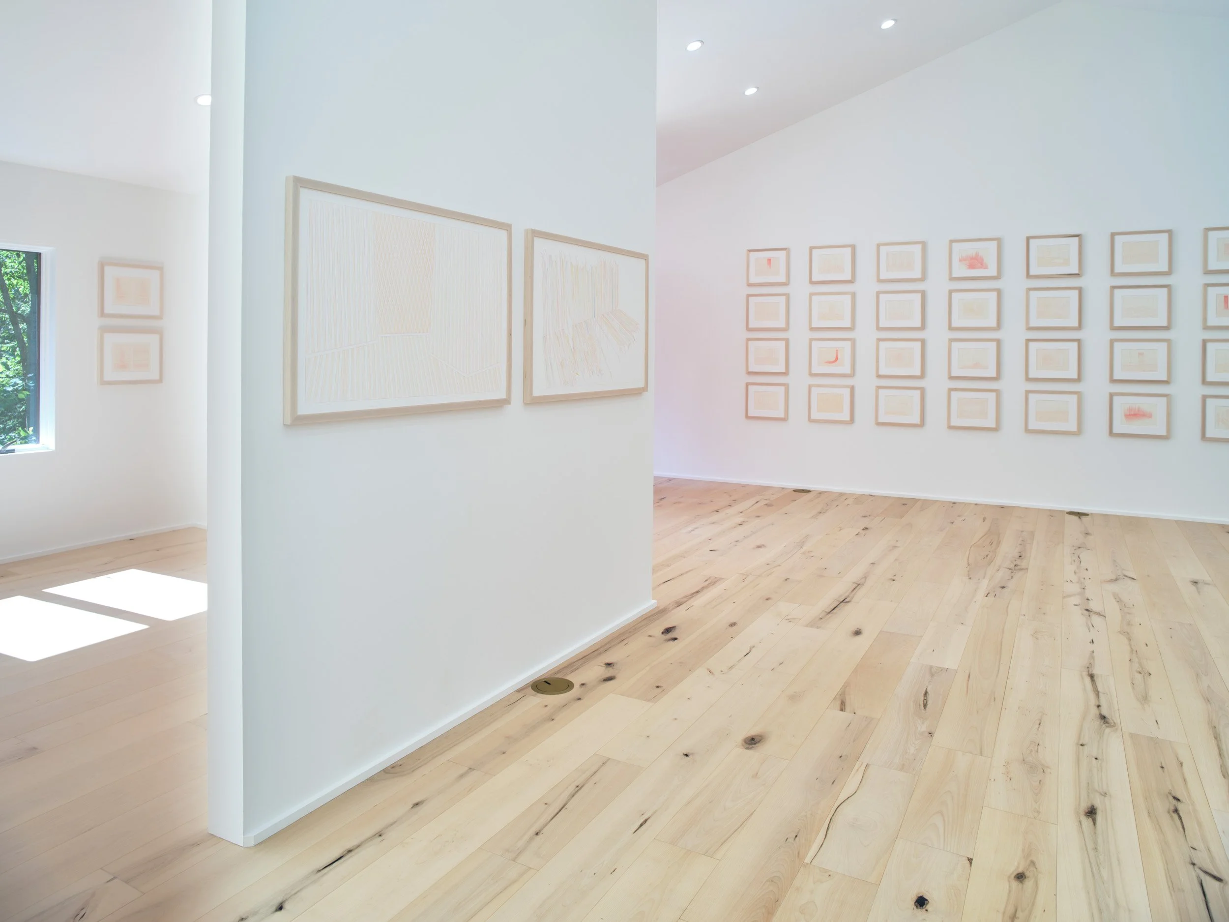An art gallery with light-colored wooden floors and white walls. Several small framed artworks are arranged on the walls, and larger pieces are displayed on the adjacent wall. Natural light comes through a window on the left, illuminating the space.