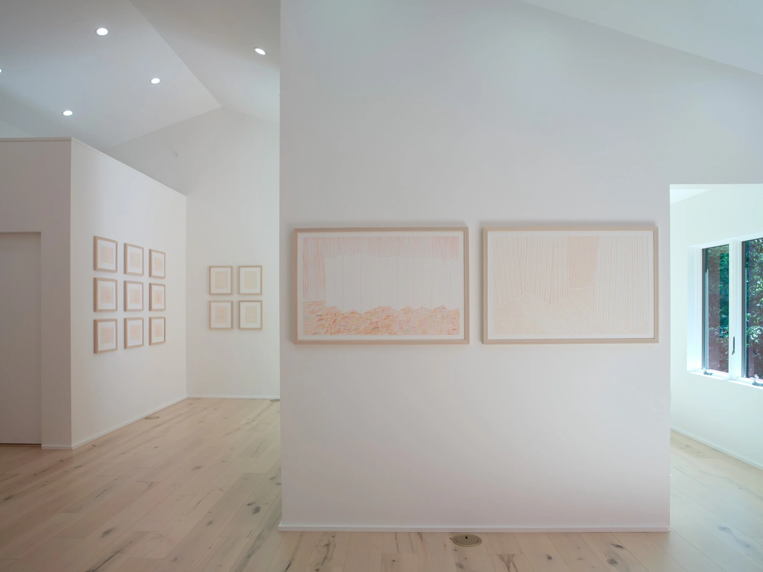 An art gallery with white walls and framed artwork, natural light from large windows, and a light-colored wooden floor.