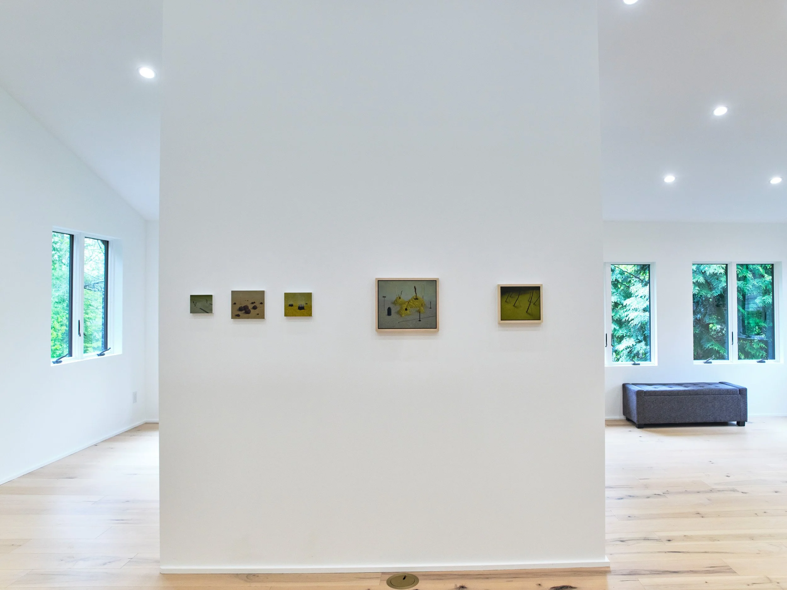 A modern white room with high ceilings, four framed pictures hanging on the main wall, two windows with green trees outside, and a gray ottoman in the corner.