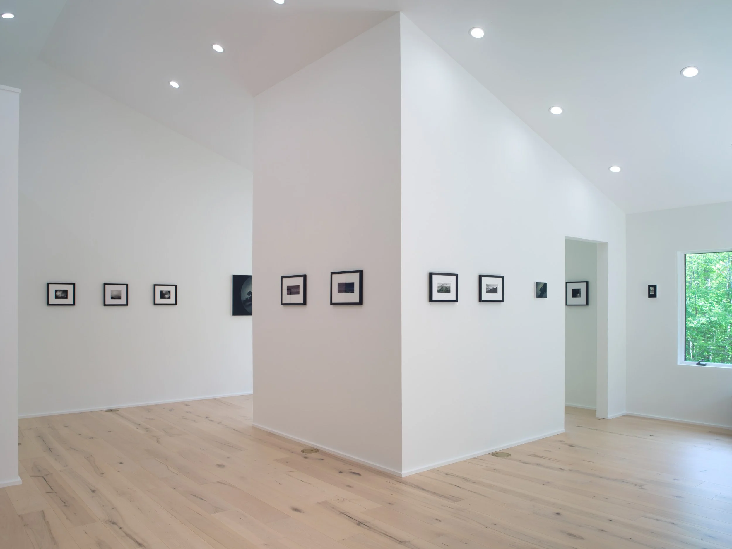 Interior view of an art gallery with white walls and wooden floors, featuring framed artwork displayed along the walls, a doorway to another room, and a large window showing greenery outside.