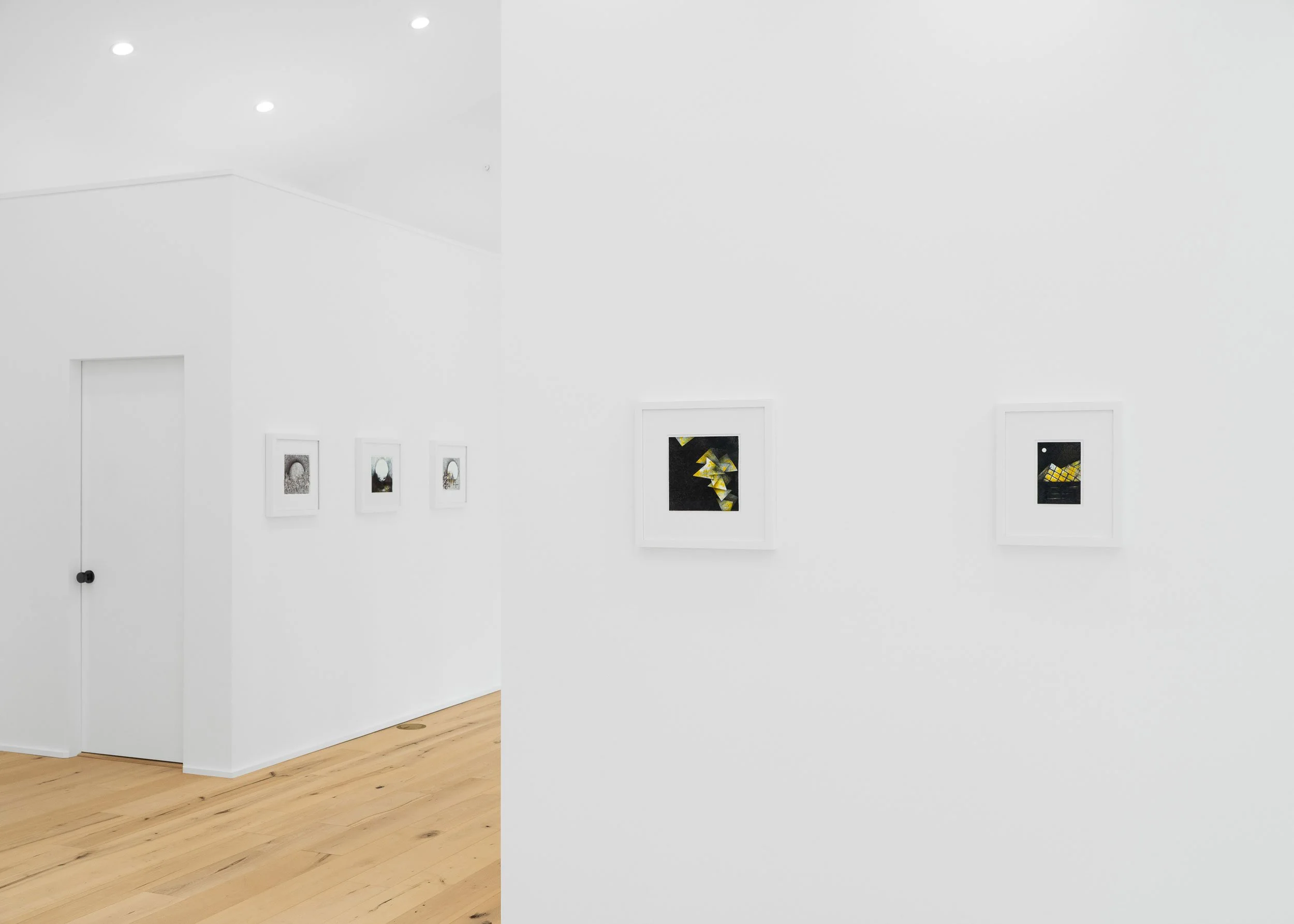 An art gallery with white walls displaying framed artwork. The room has a light wooden floor and recessed ceiling lights.