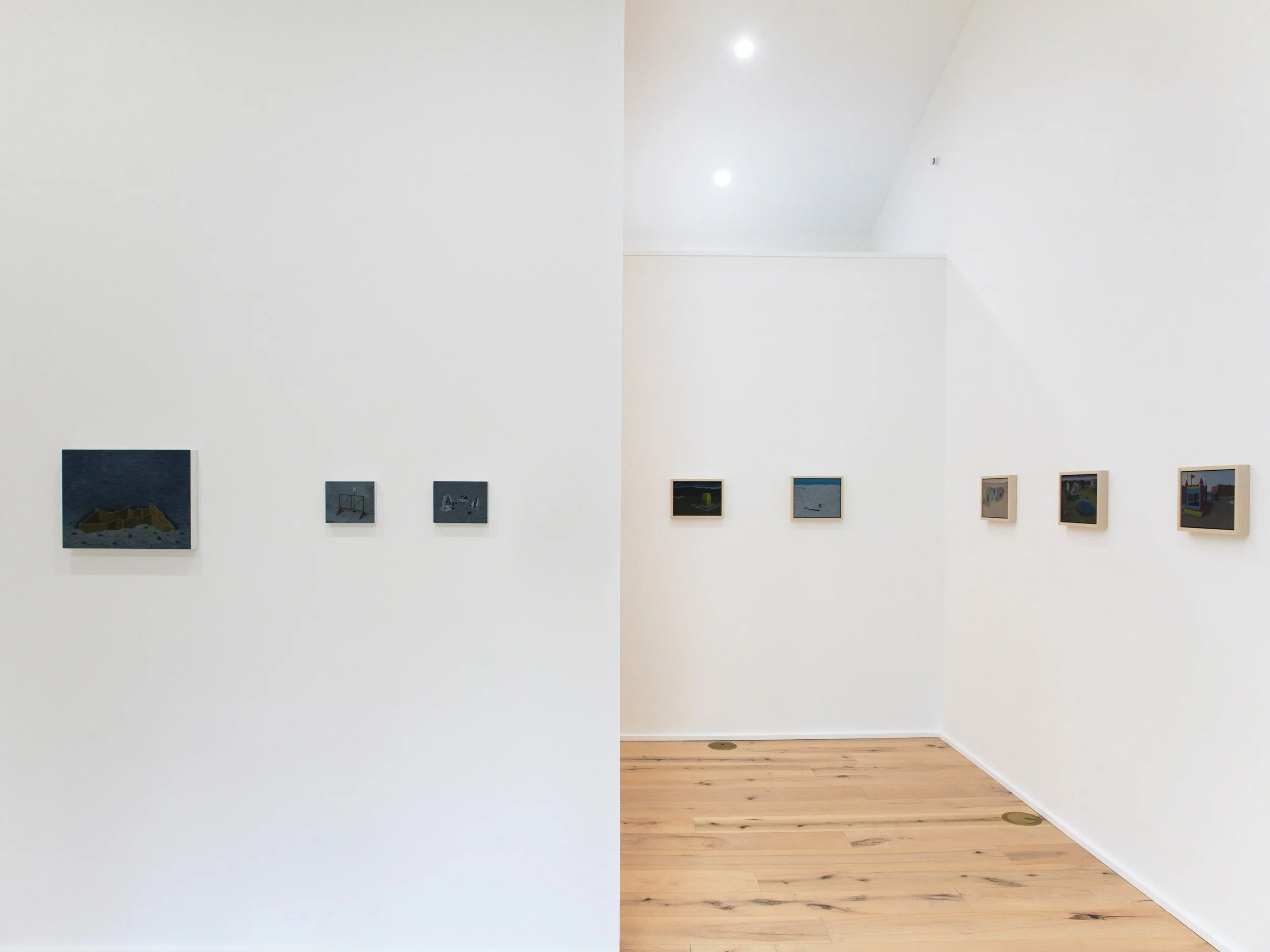 Photograph of an art gallery with plain white walls and a wooden floor, featuring small framed artworks evenly spaced along the walls, under bright ceiling lights.