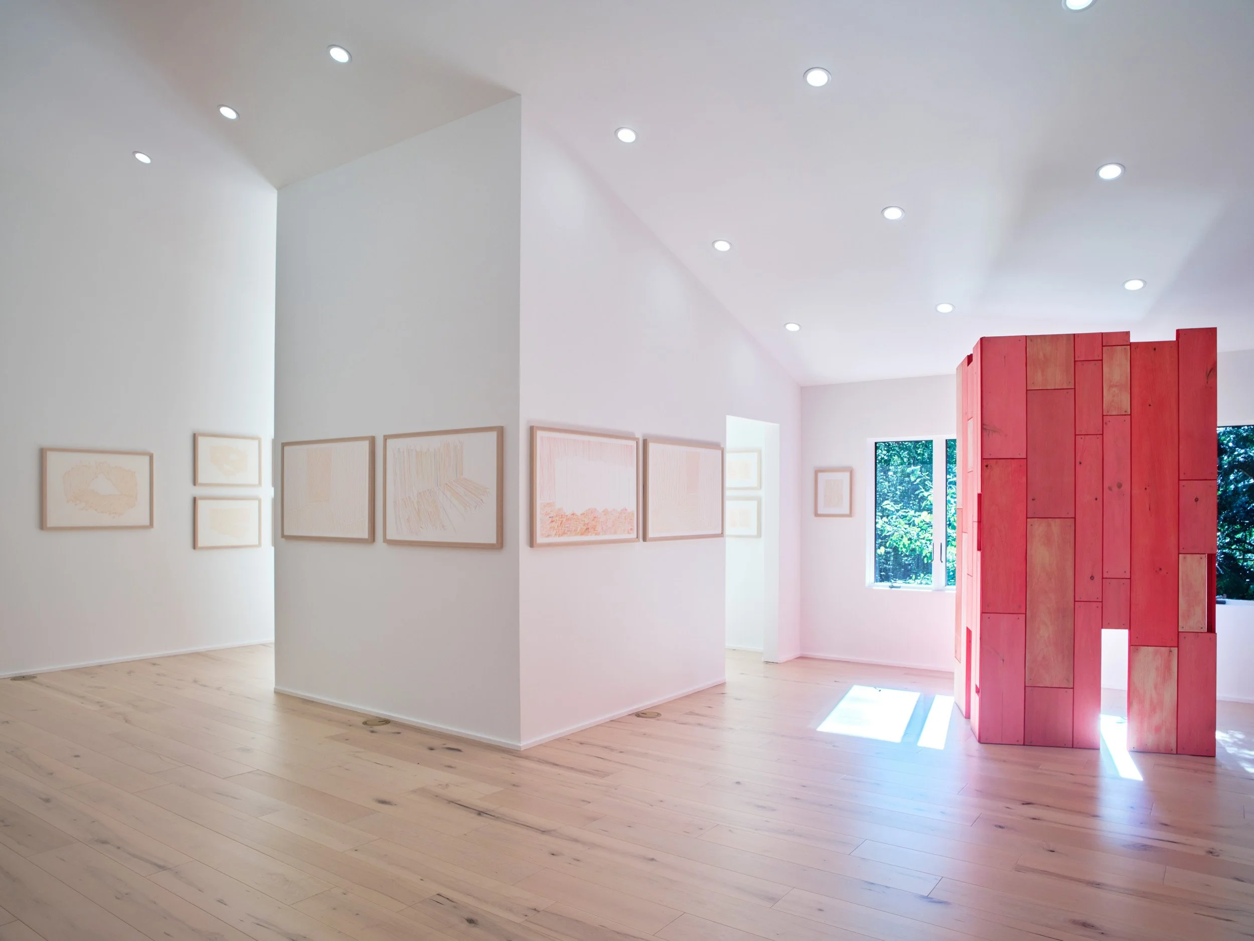 A contemporary art gallery with white walls and light wooden floors, displaying framed artwork and a pinkish-red wooden sculpture near a window.