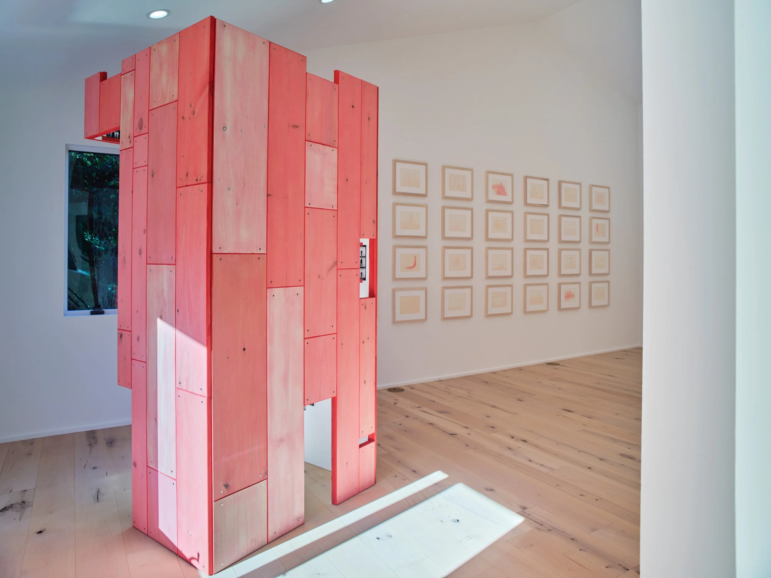 An art installation featuring a large structure made of pink-painted wooden panels in a gallery. The structure extends upward and outward, with a small window on the left side. Behind it, beige framed pictures are arranged in a grid on a white gallery wall. The gallery has light-colored hardwood floors and a single window through which trees can be seen.