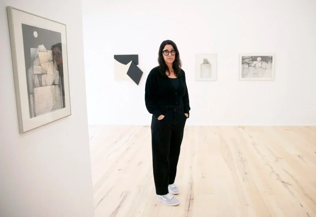 A woman wearing glasses, black jacket, black pants, and sneakers standing in an art gallery with framed artworks on white walls and light-colored flooring.