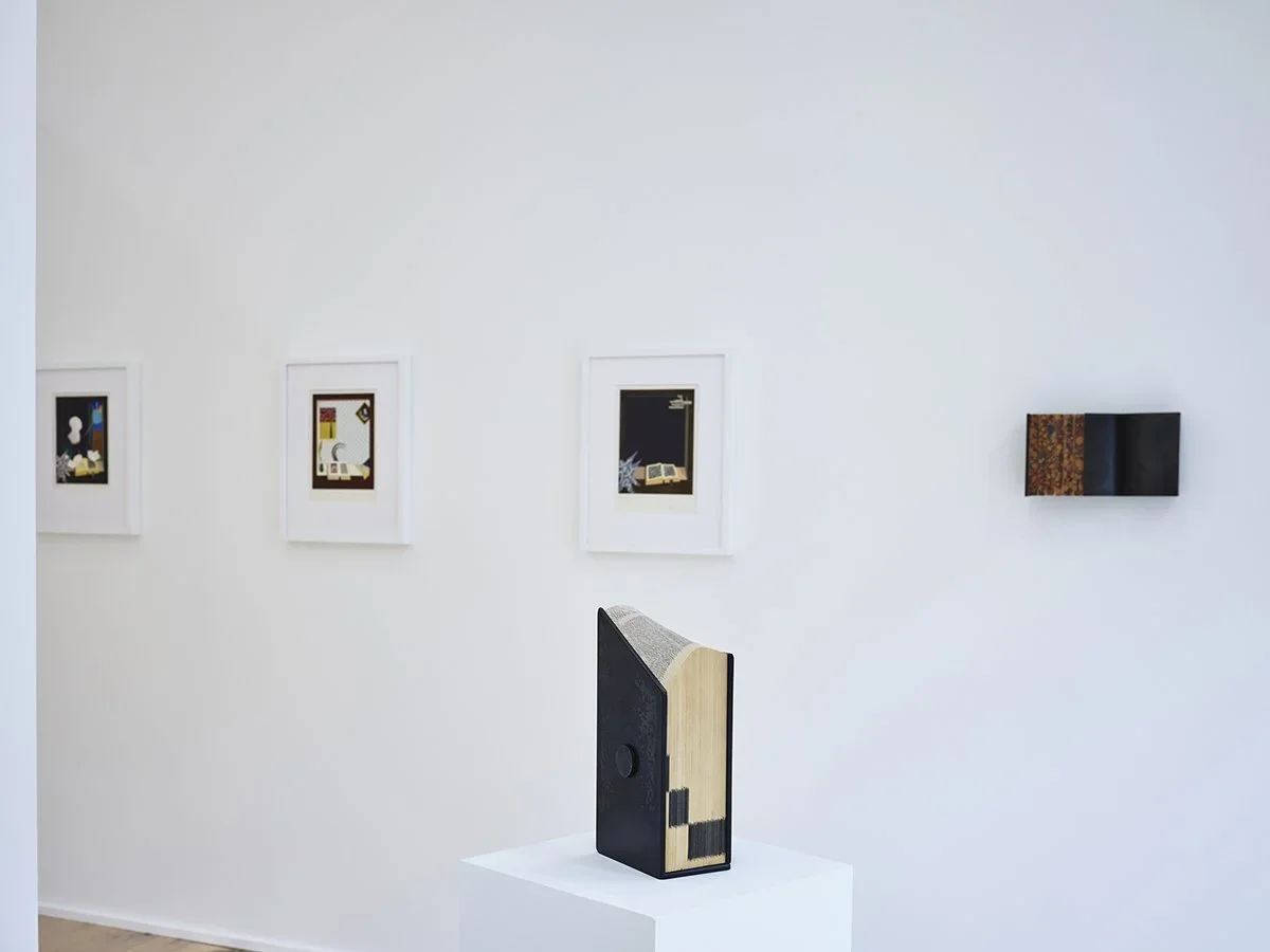 Gallery with artworks, including framed pictures on the wall and a sculptural book on a pedestal.