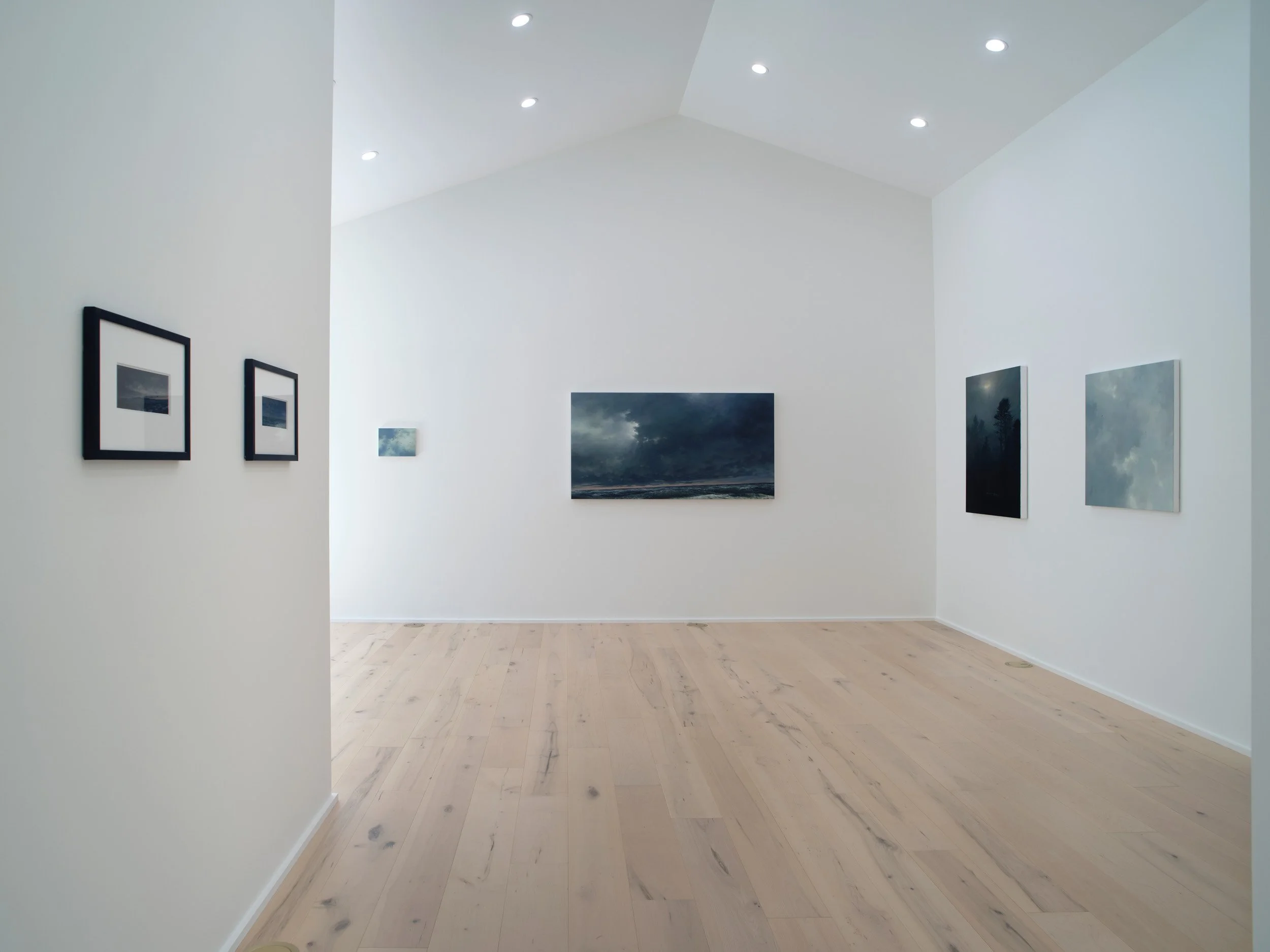 A corner of an art gallery with white walls, light wood flooring, and framed photographs and paintings on the walls.