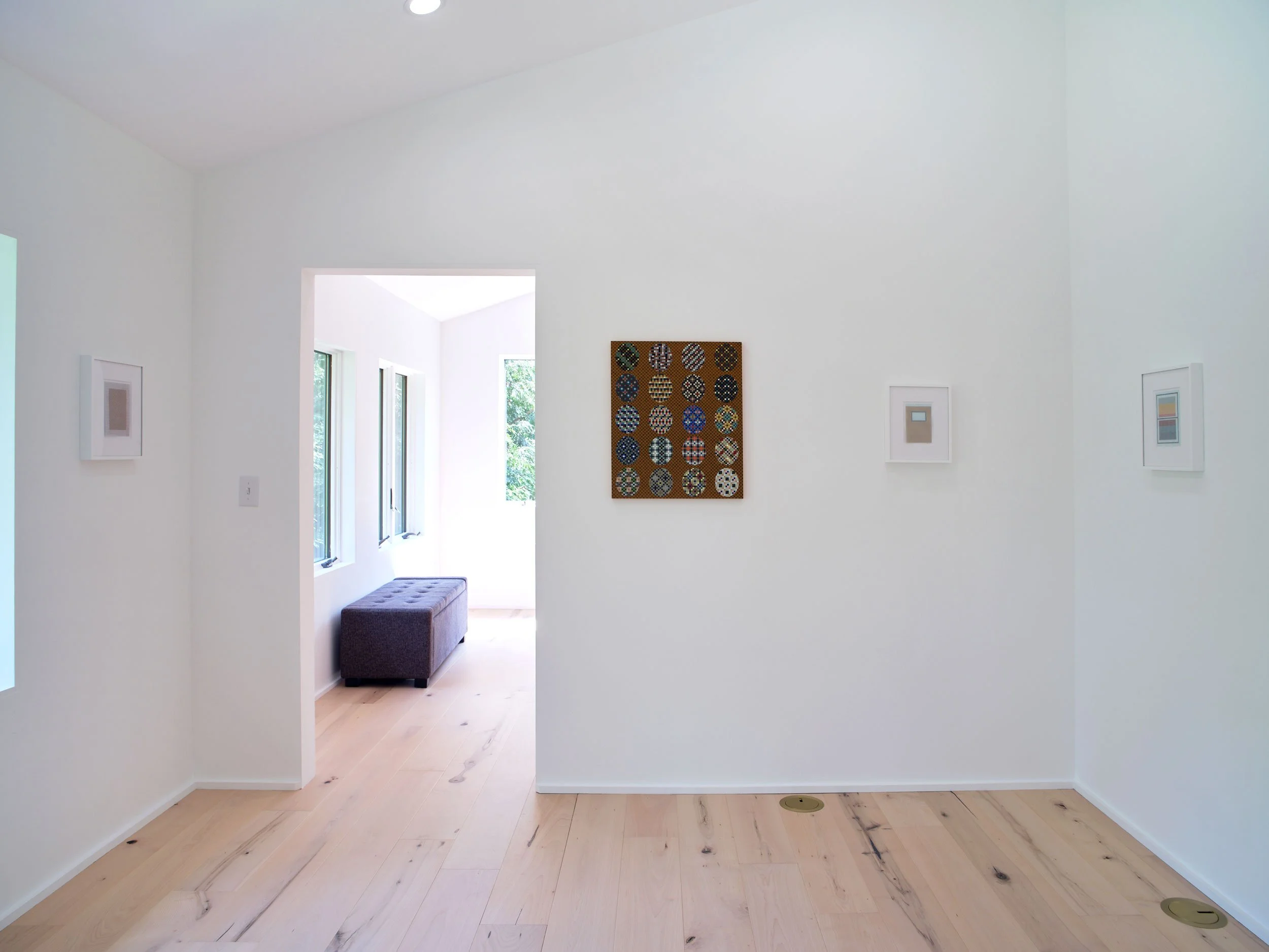 Interior view of a bright, white room with hardwood floors, a doorway leading to an outdoor area with trees, and artwork on the walls.