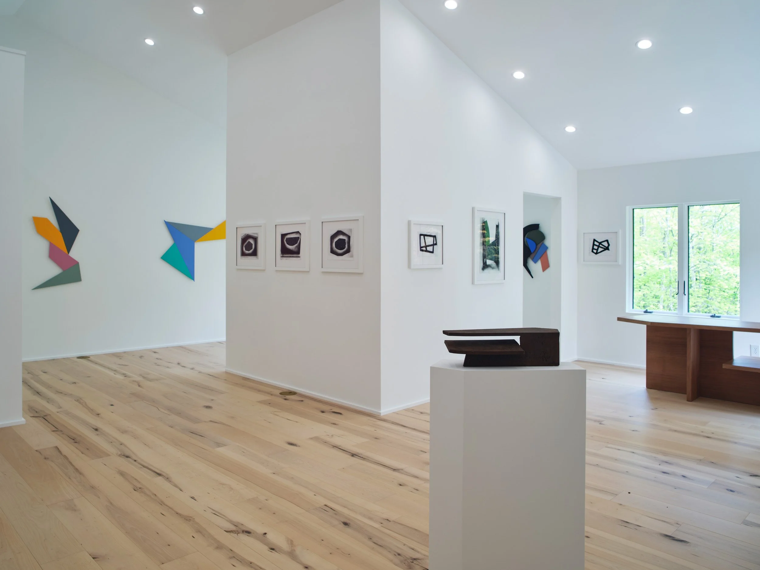 Art gallery with abstract and modern artworks displayed on white walls, natural light from windows, and wooden flooring.