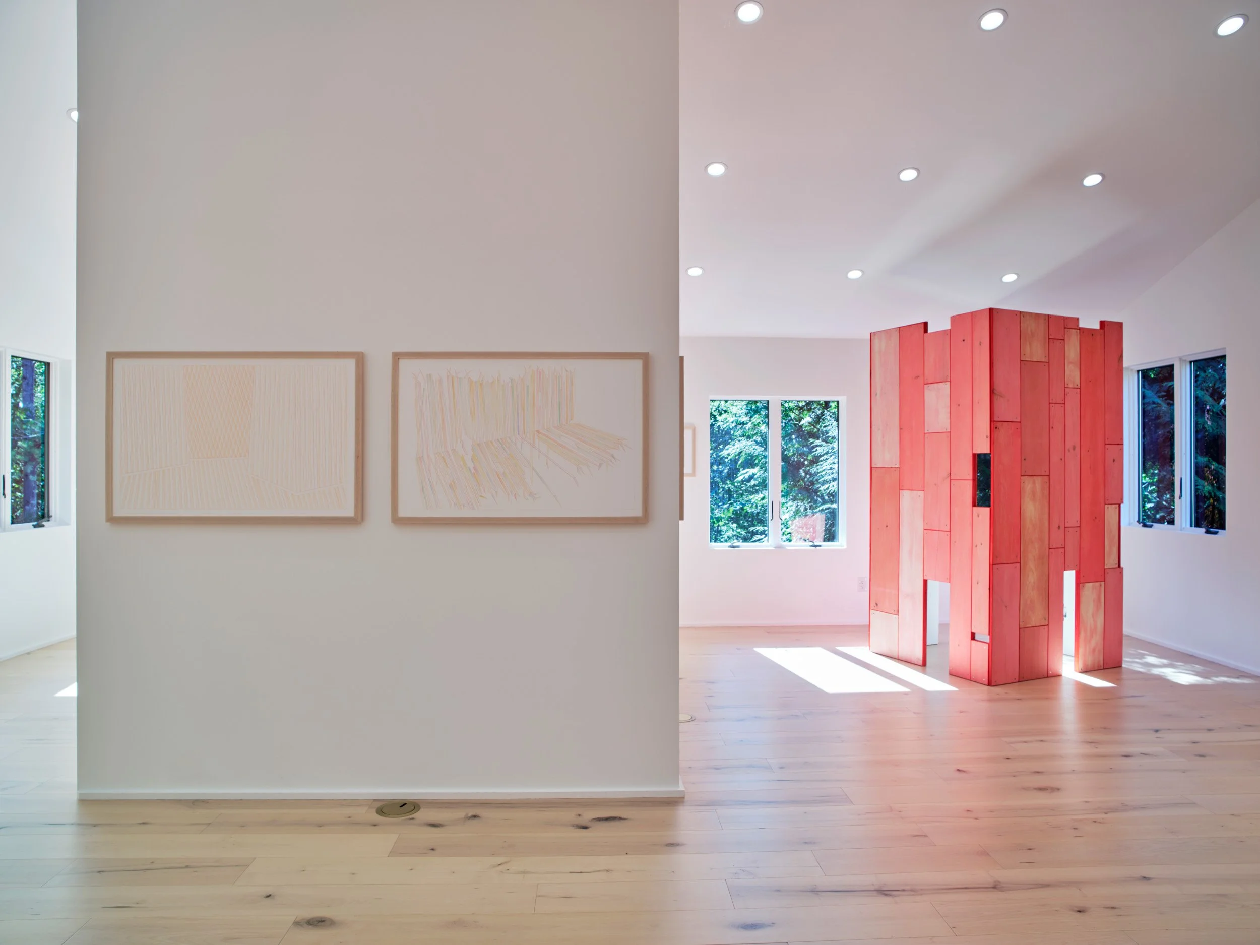 Interior view of an art gallery with white walls, wooden flooring, and natural light from windows. Artwork is displayed on the wall, and there is a red wooden sculpture in the background.