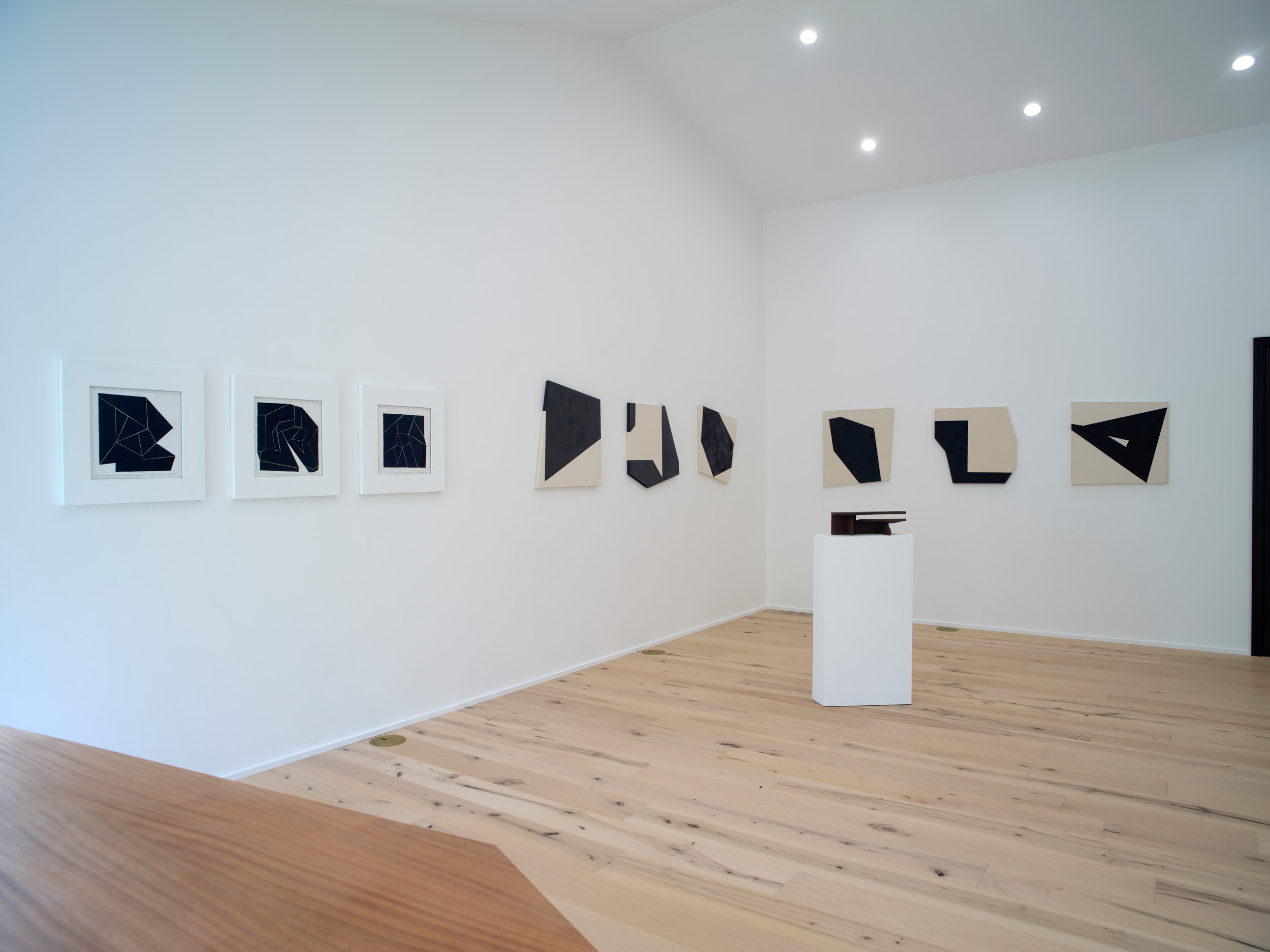 Gallery with framed abstract black and white artwork on a white wall, and a small sculpture on a white pedestal, in a minimalistic art space with wood flooring and bright ceiling lights.