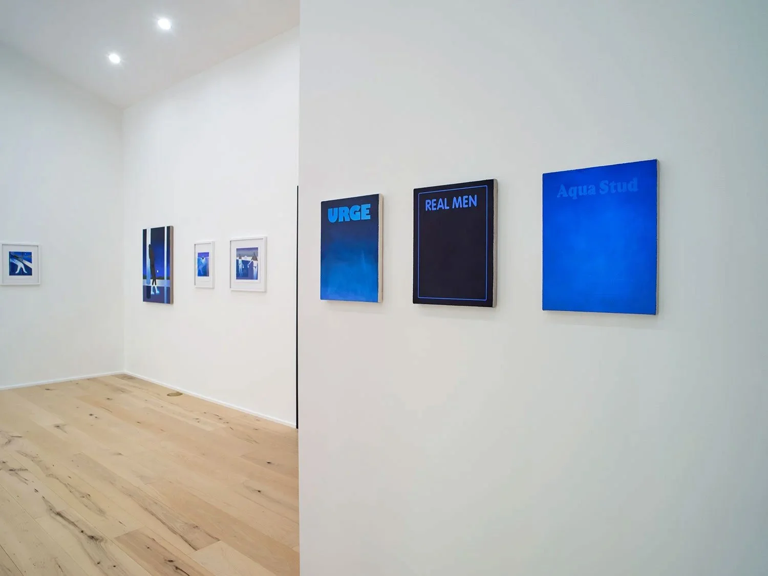 Art gallery with multiple blue and black paintings, some with text like 'URGE,' 'REAL MEN,' and 'Aqua Stud,' displayed on white walls and wooden floors.