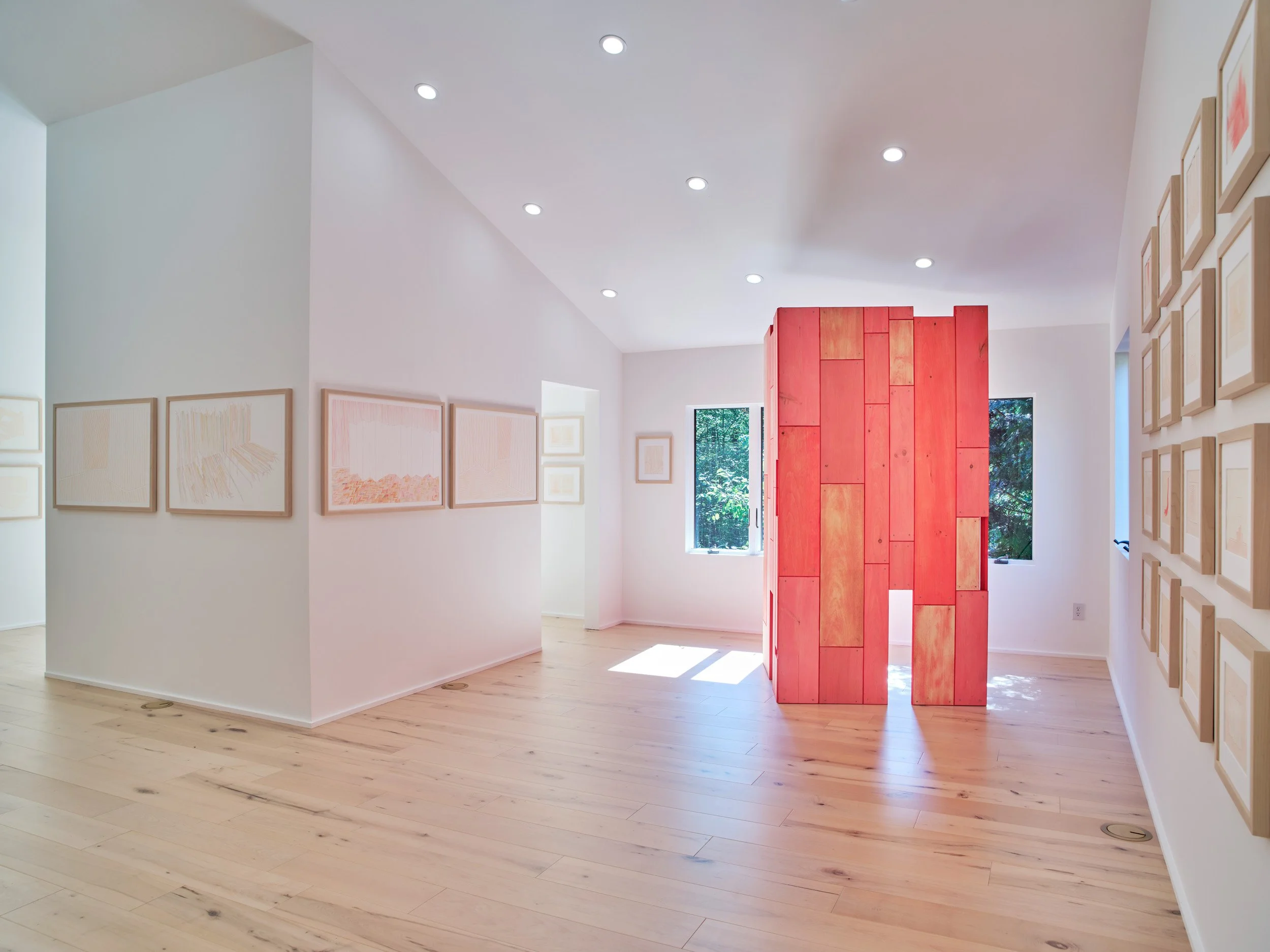 An art gallery with white walls and light wood floors, featuring multiple framed artworks on the walls and a central red art piece made of stacked wooden panels, with windows allowing natural light.