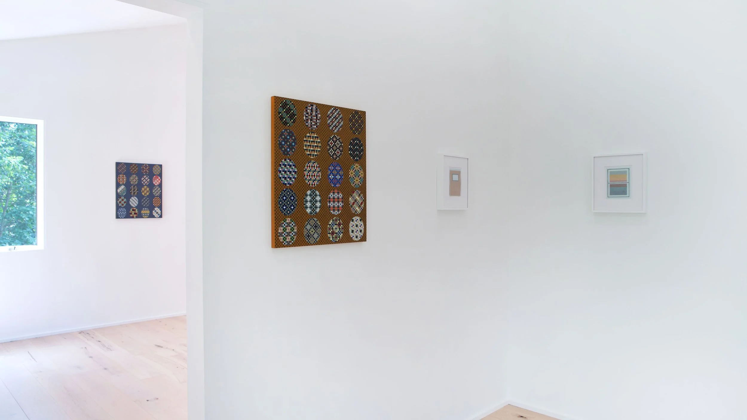Interior of an art gallery with white walls, displaying framed modern artwork including a large piece with patterned circles, a smaller artwork with horizontal color blocks, and another with small shapes, natural light from window, light wood floor.