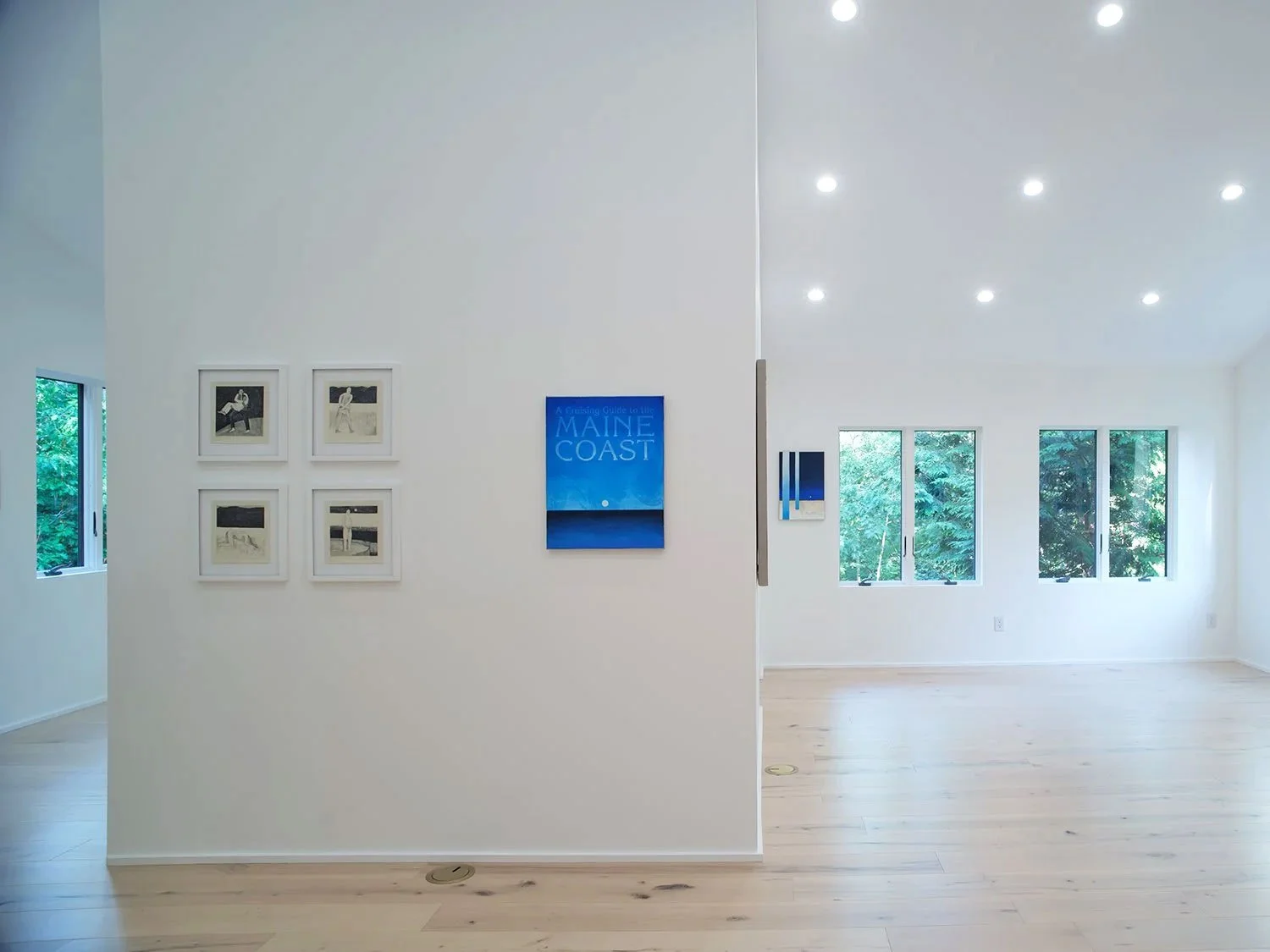 An art gallery with white walls, displaying framed artwork and a large blue book cover titled "A Cruising Guide to the Maine Coast." There are 4 small black-and-white sketches on the left wall and three windows on the right side with lush green trees outside. The ceiling has recessed lighting, and the floor is light wood.