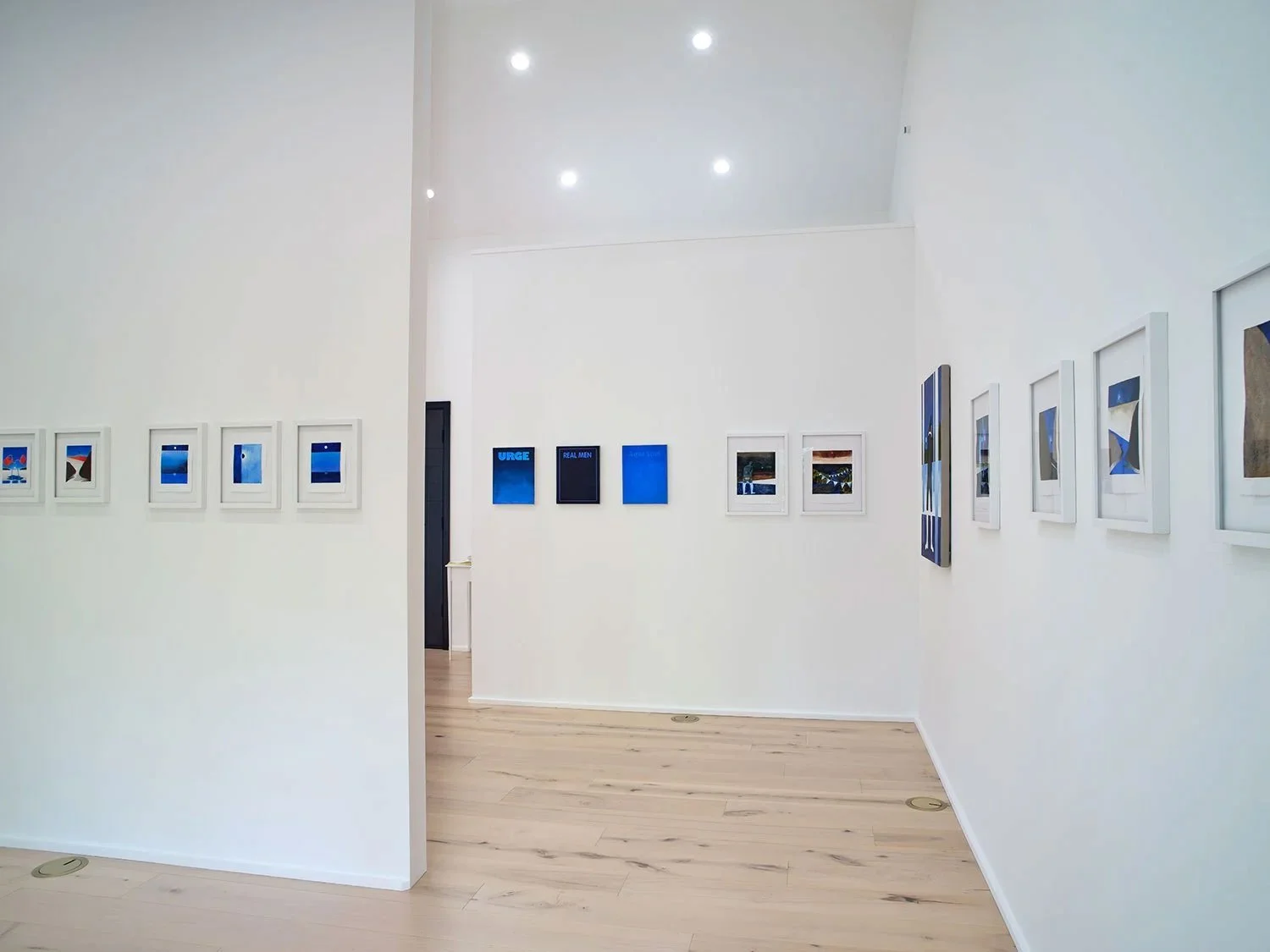 Gallery with white walls displaying framed artworks and blue-themed posters, wooden floor, ceiling spotlights.