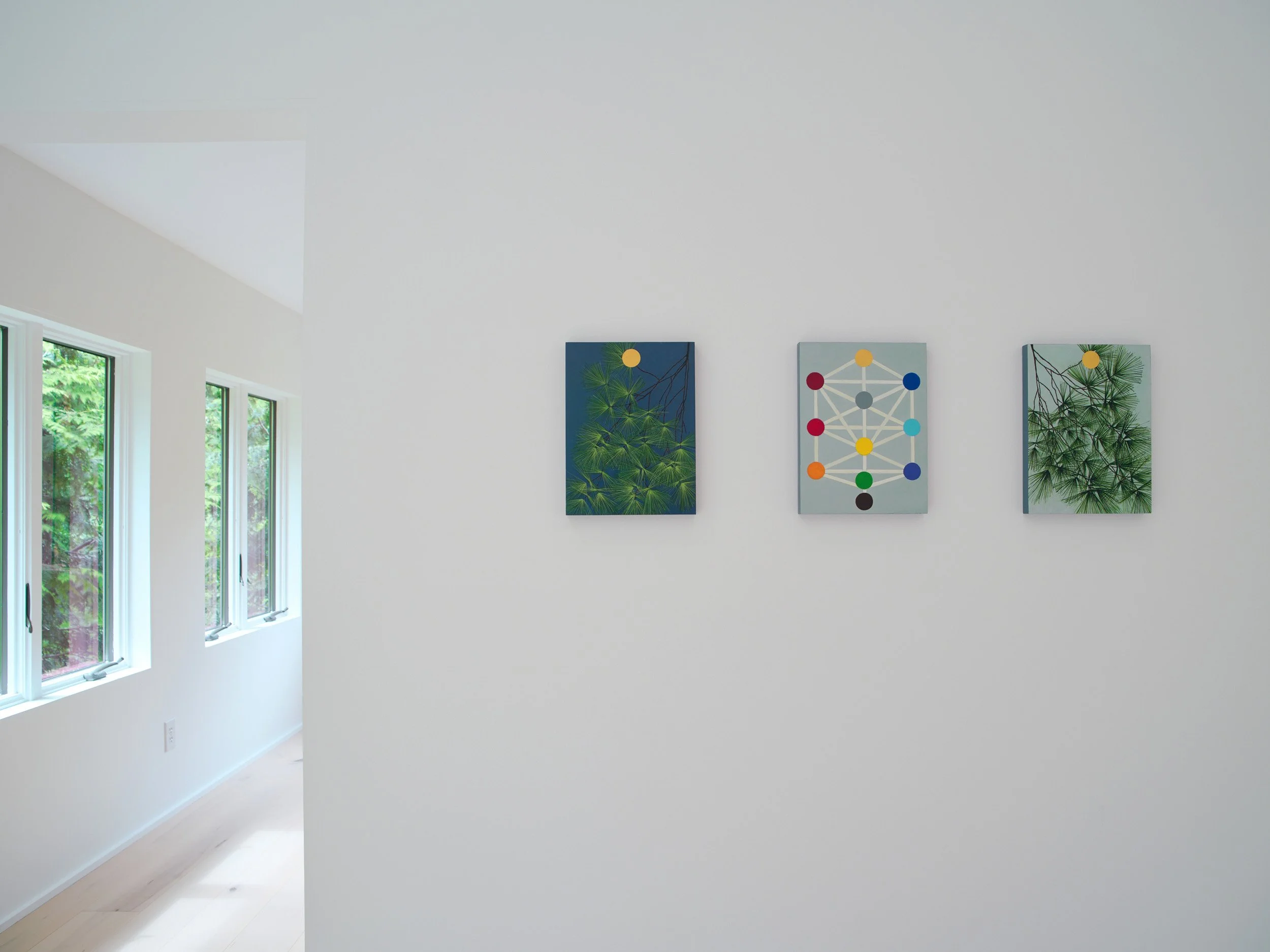 White gallery wall with three colorful modern art paintings, two side windows show greenery outside.
