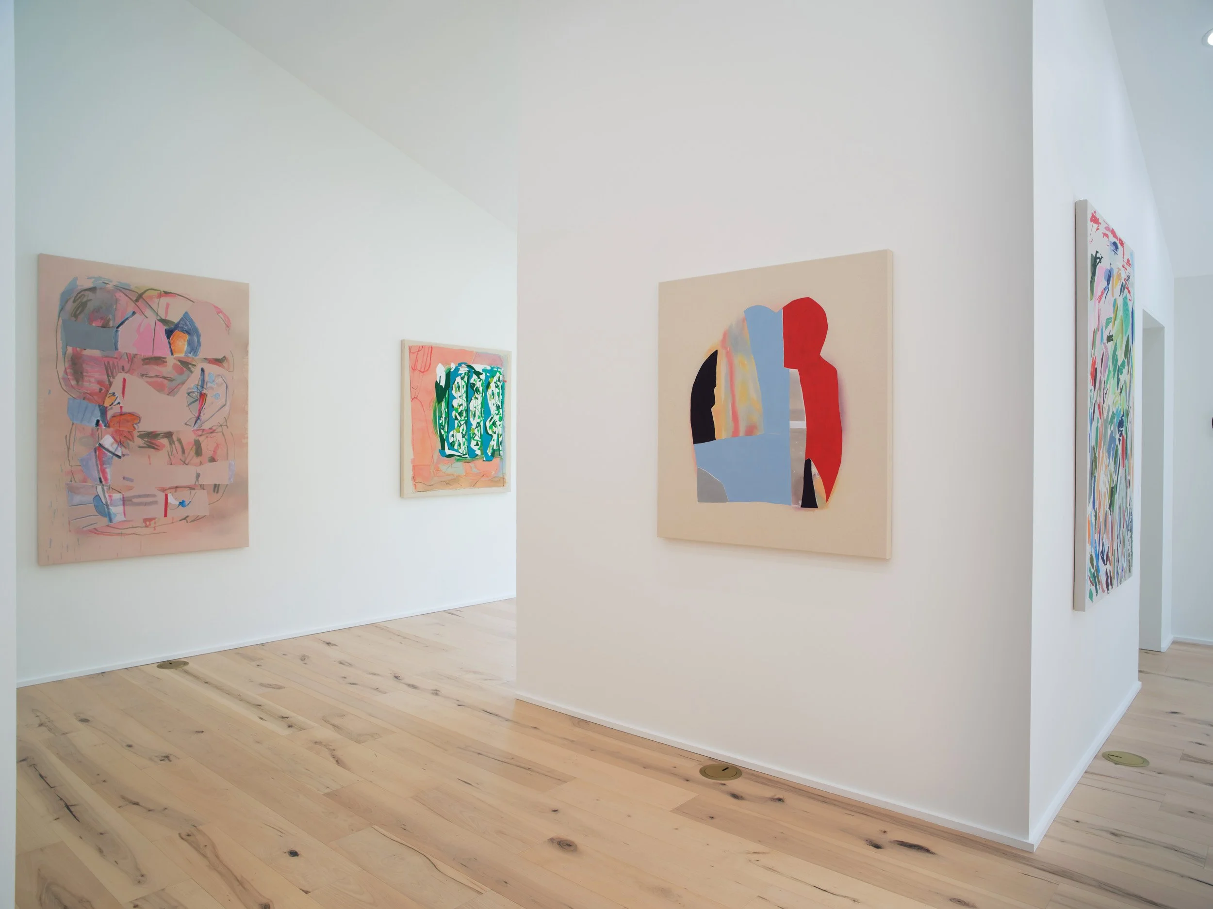 View of an art gallery with white walls displaying colorful abstract paintings, wooden flooring, and partial view of a doorway on the right side.
