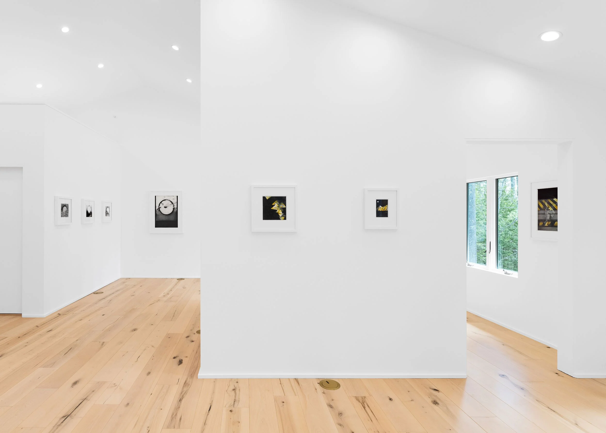 A modern art gallery with white walls, light-colored wooden floors, and framed artwork hanging on the walls. There are windows letting in natural light.