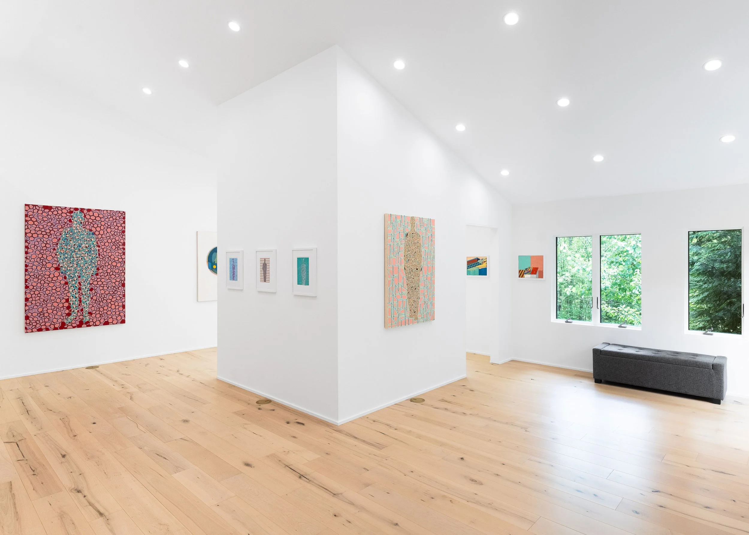 Interior of an art gallery with white walls, hardwood floors, several colorful paintings, large windows showing green trees, and a charcoal sofa.