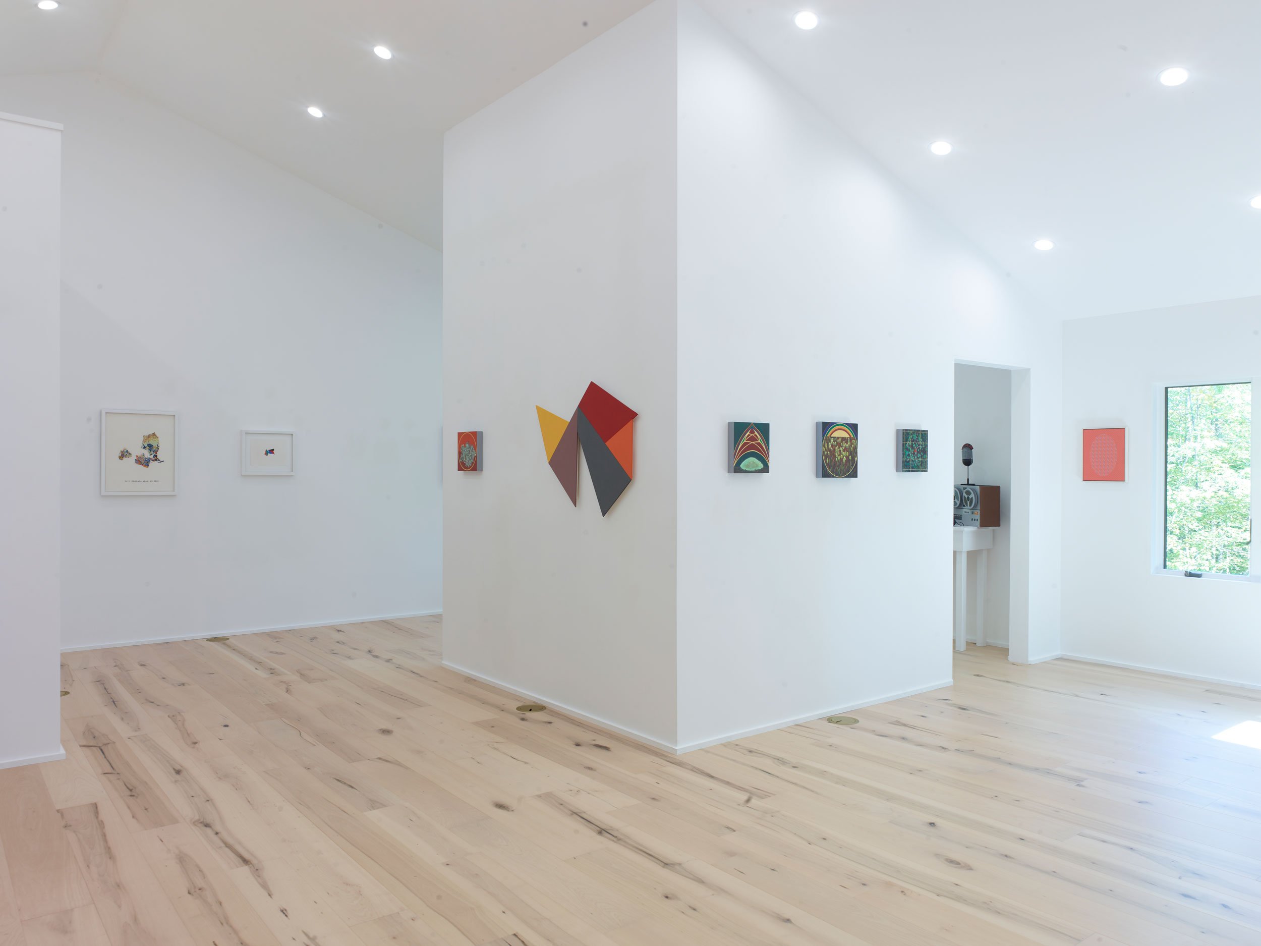 Interior of an art gallery with white walls and wooden floors, displaying various colorful artworks. Natural light is coming in through large windows.