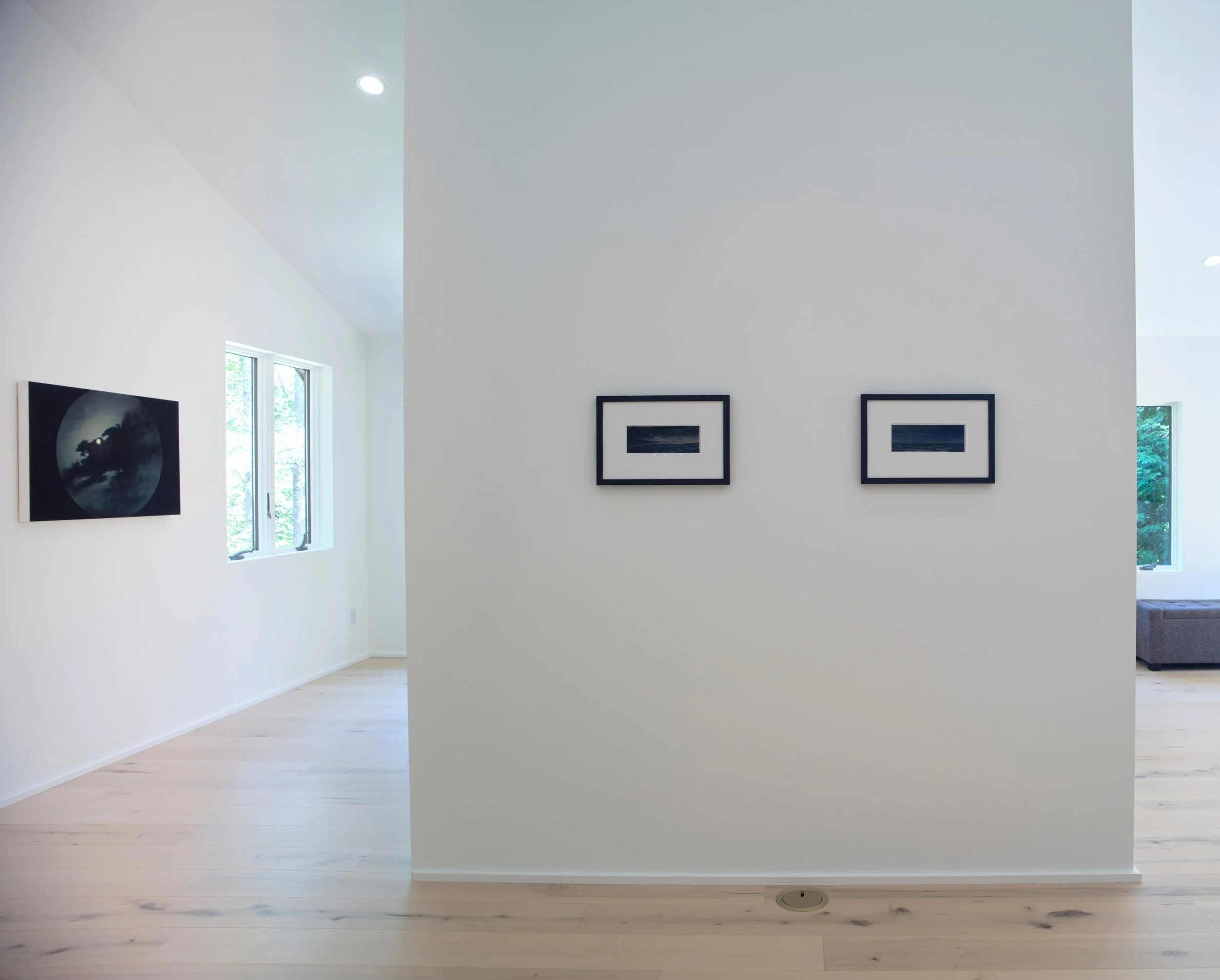 A modern, minimalistic art gallery with white walls, light wooden flooring, and framed artwork. There are windows and a glimpse of a gray sofa in the background.