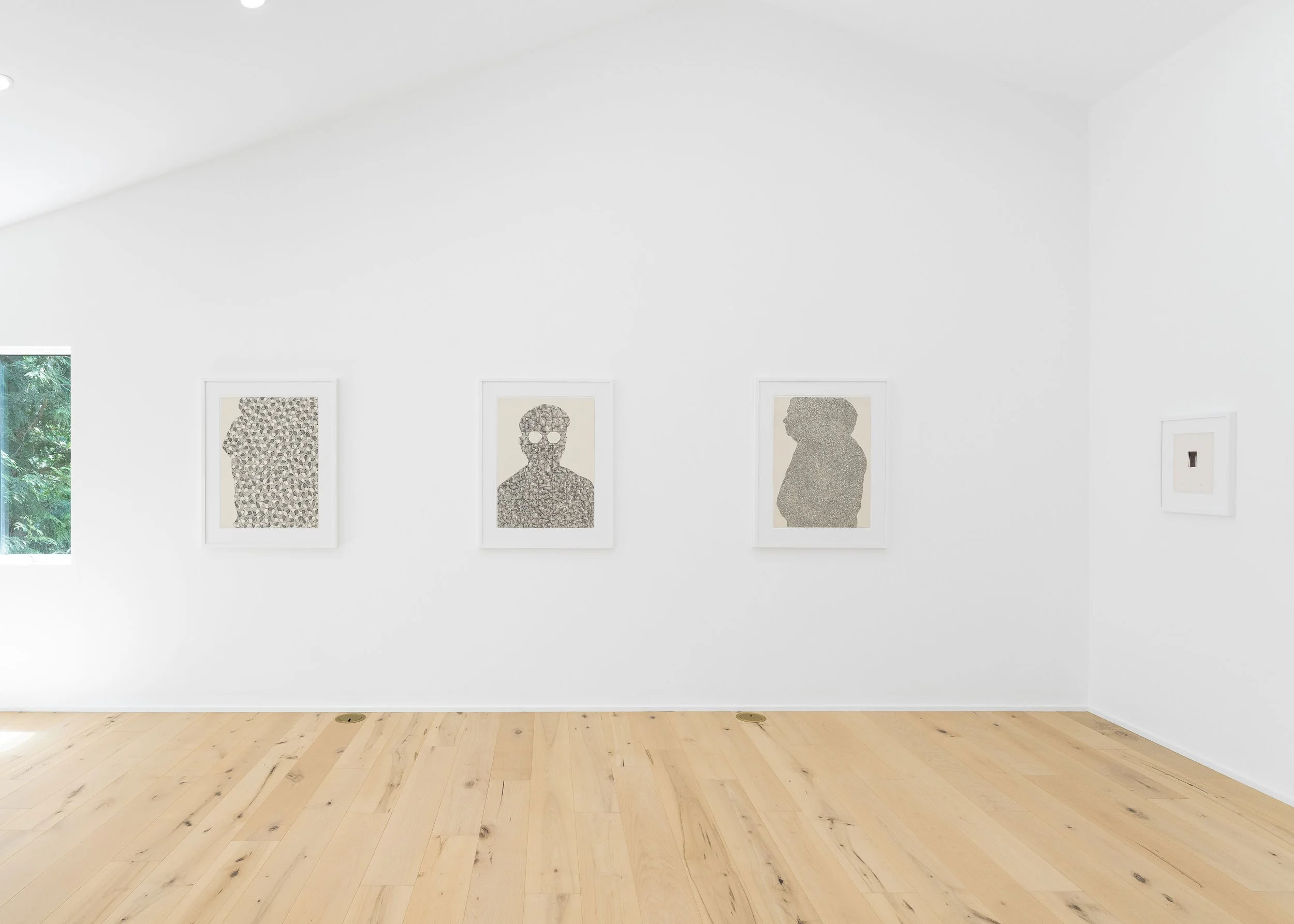 Gallery wall with four framed portraits of human figures, with a window showing greenery on the left; wooden floor; white walls.